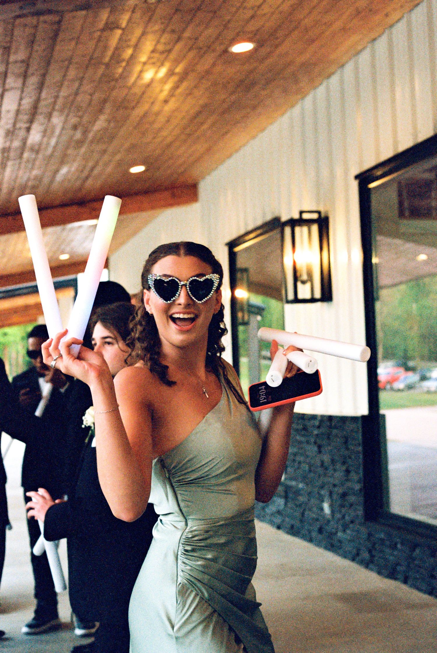 Bridesmaid in sage green dress wearing heart sunglasses holding foam sticks at wedding.