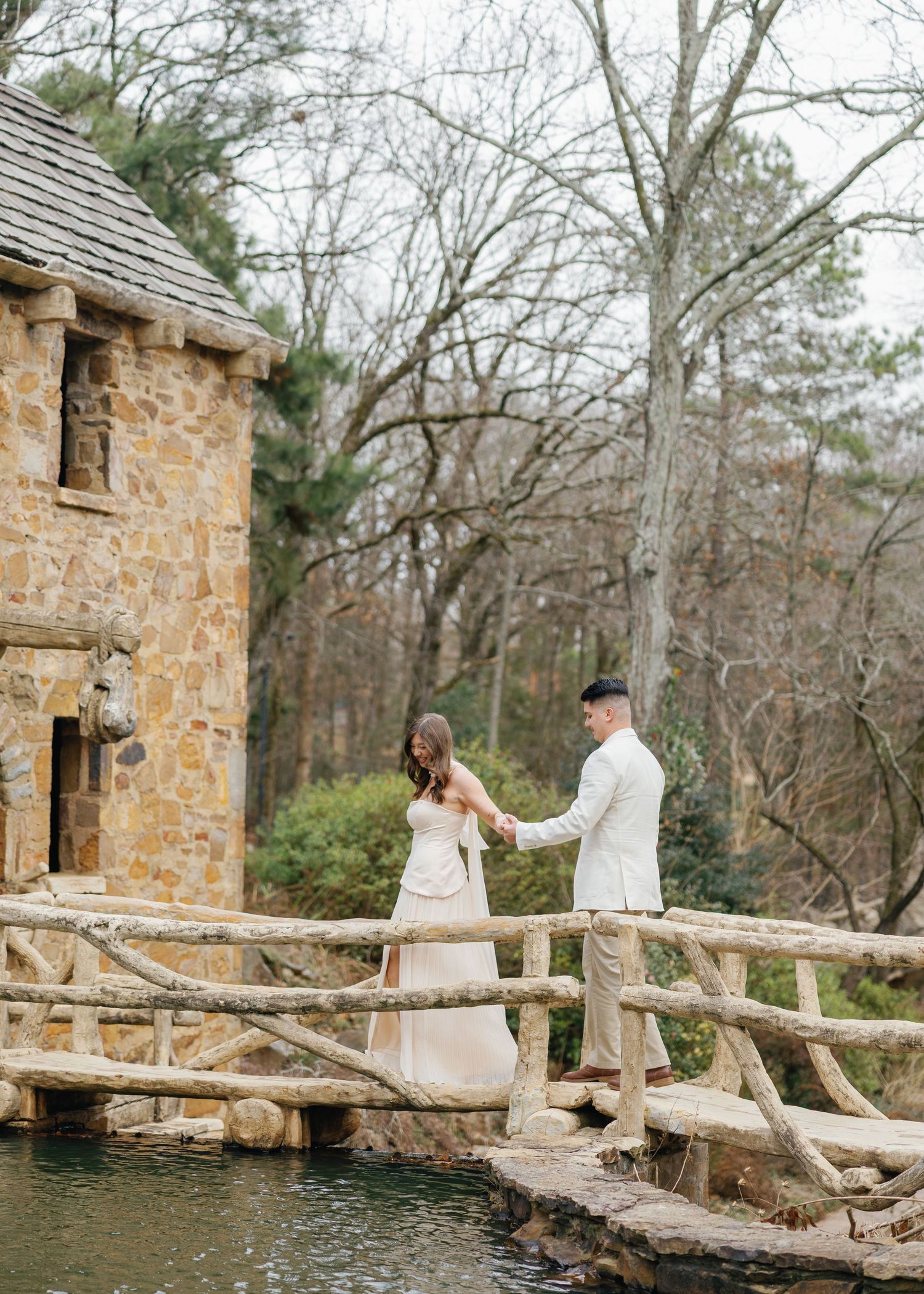 Couple holding hands on a rustic wooden bridge beside a stone building near a serene pond.