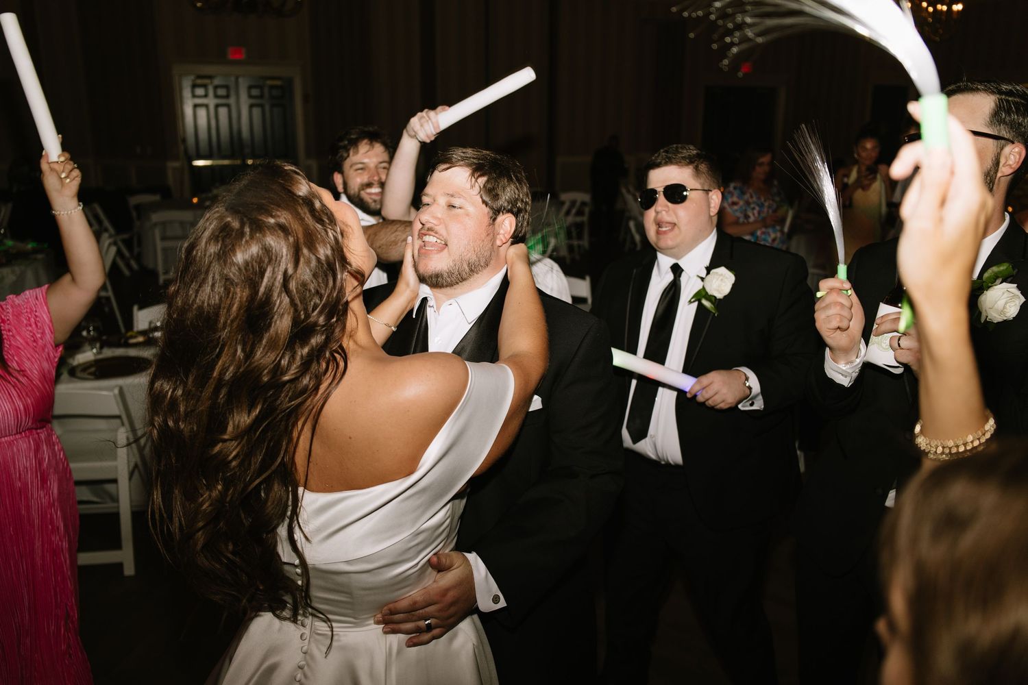 Bride and groom dance joyfully with guests waving glow sticks at lively wedding reception celebration.