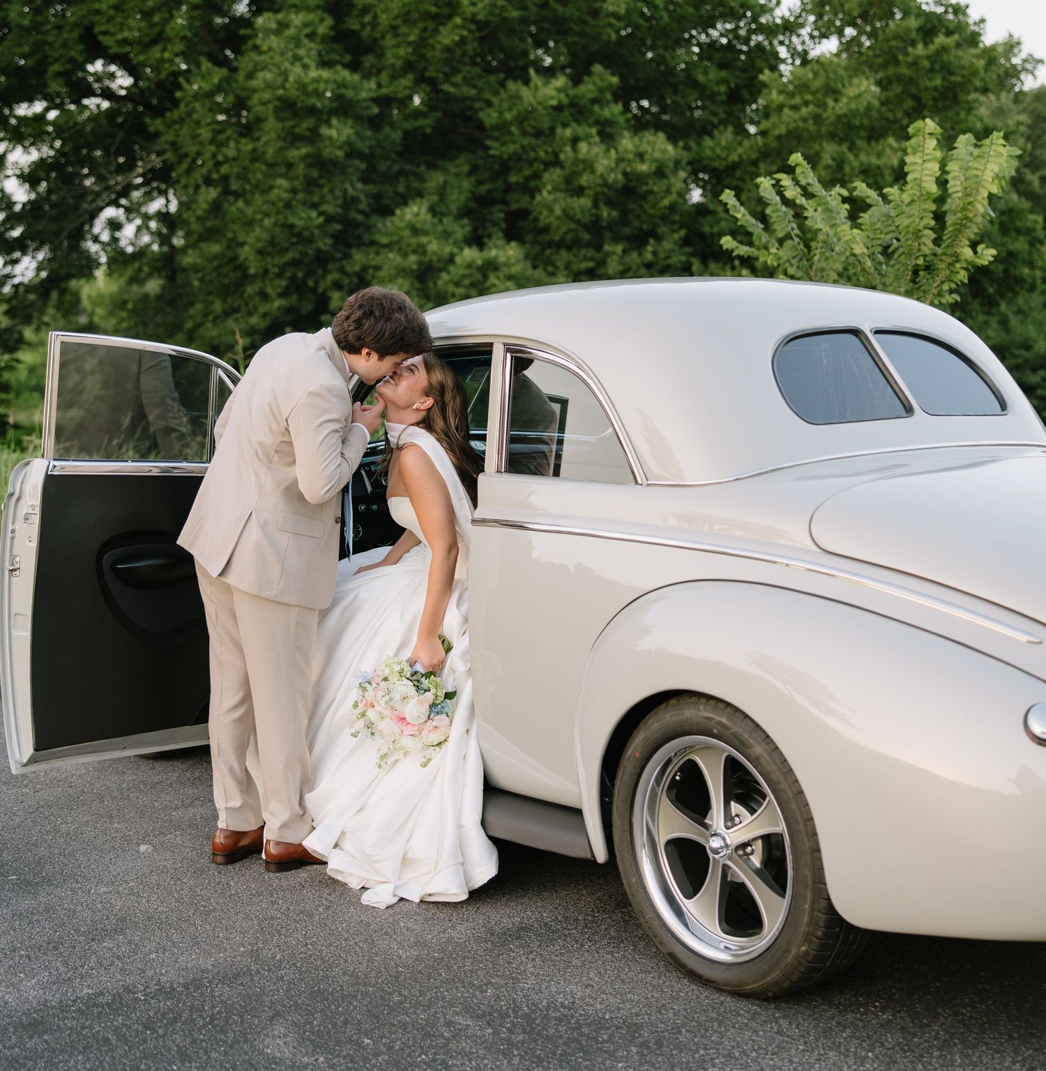 Bride and groom sharing a kiss beside a classic white vintage car on their wedding day.