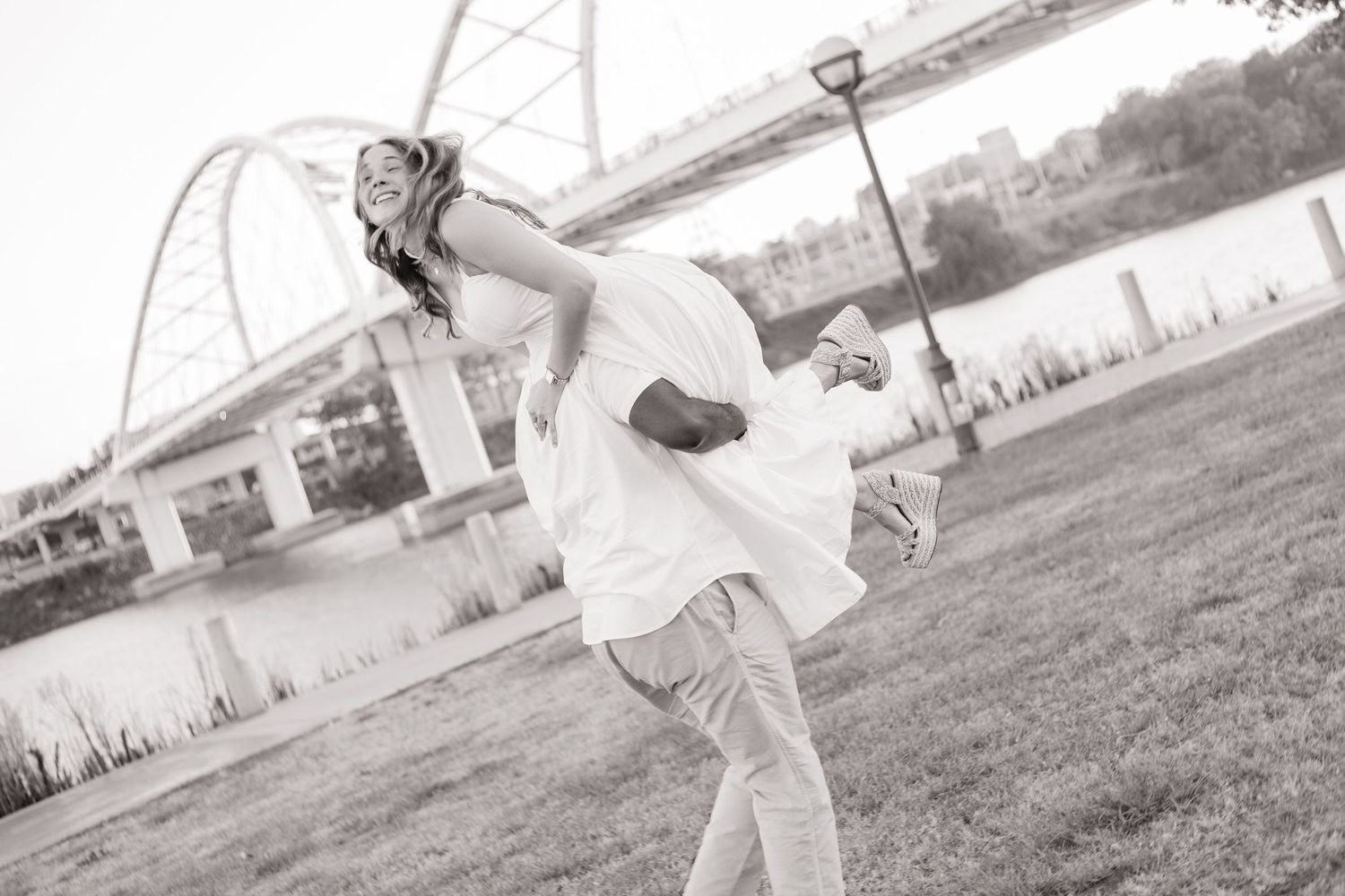 Black and white photo of groom lifting bride outdoors near a bridge, celebrating their wedding day.