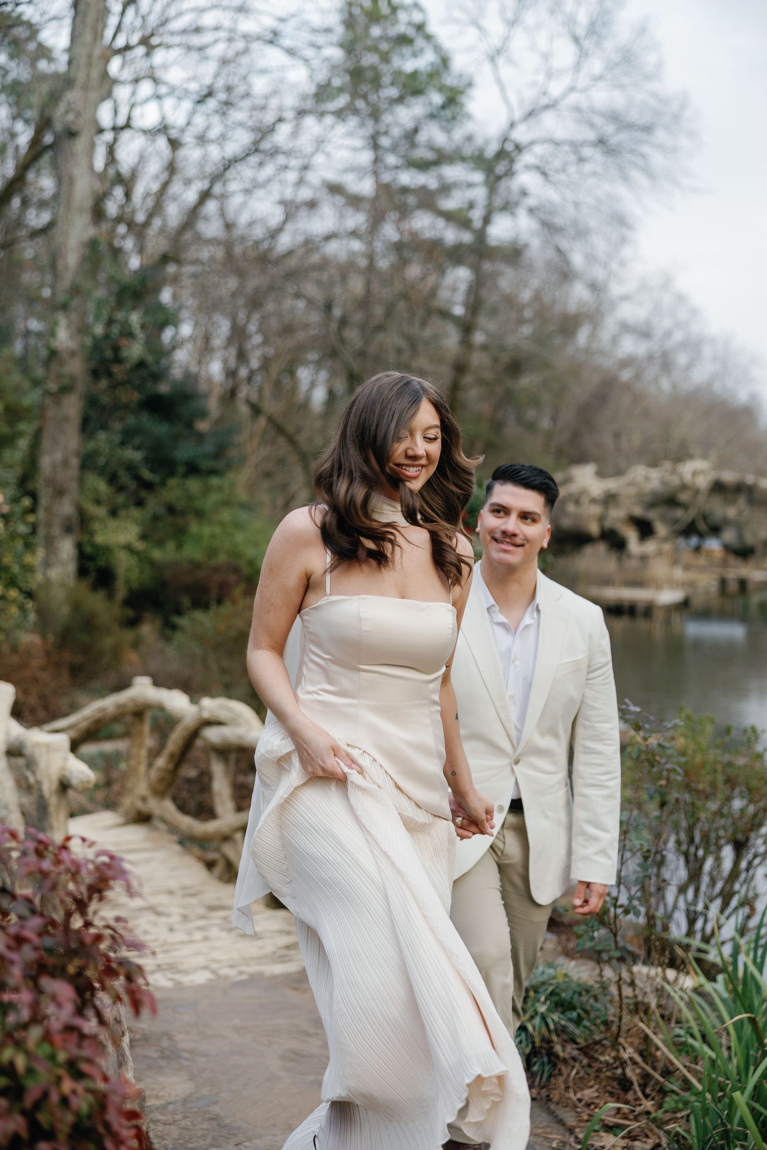 Couple in elegant cream attire walking hand-in-hand along a garden path near a pond, surrounded by lush greenery.