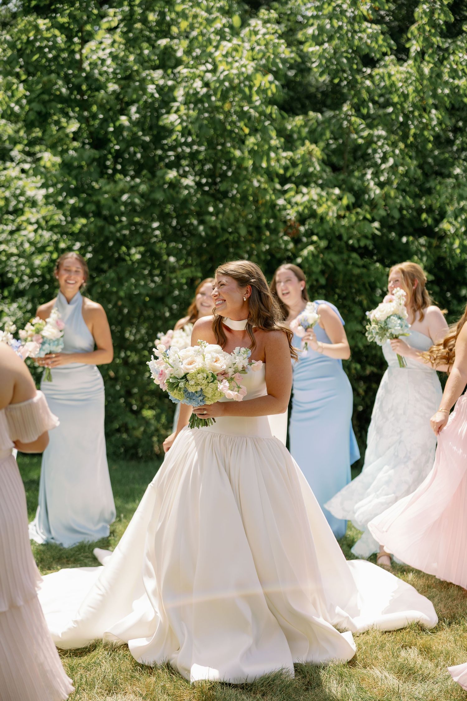 Bride in white ball gown holds floral bouquet surrounded by bridesmaids in light blue dresses on sunny garden lawn.