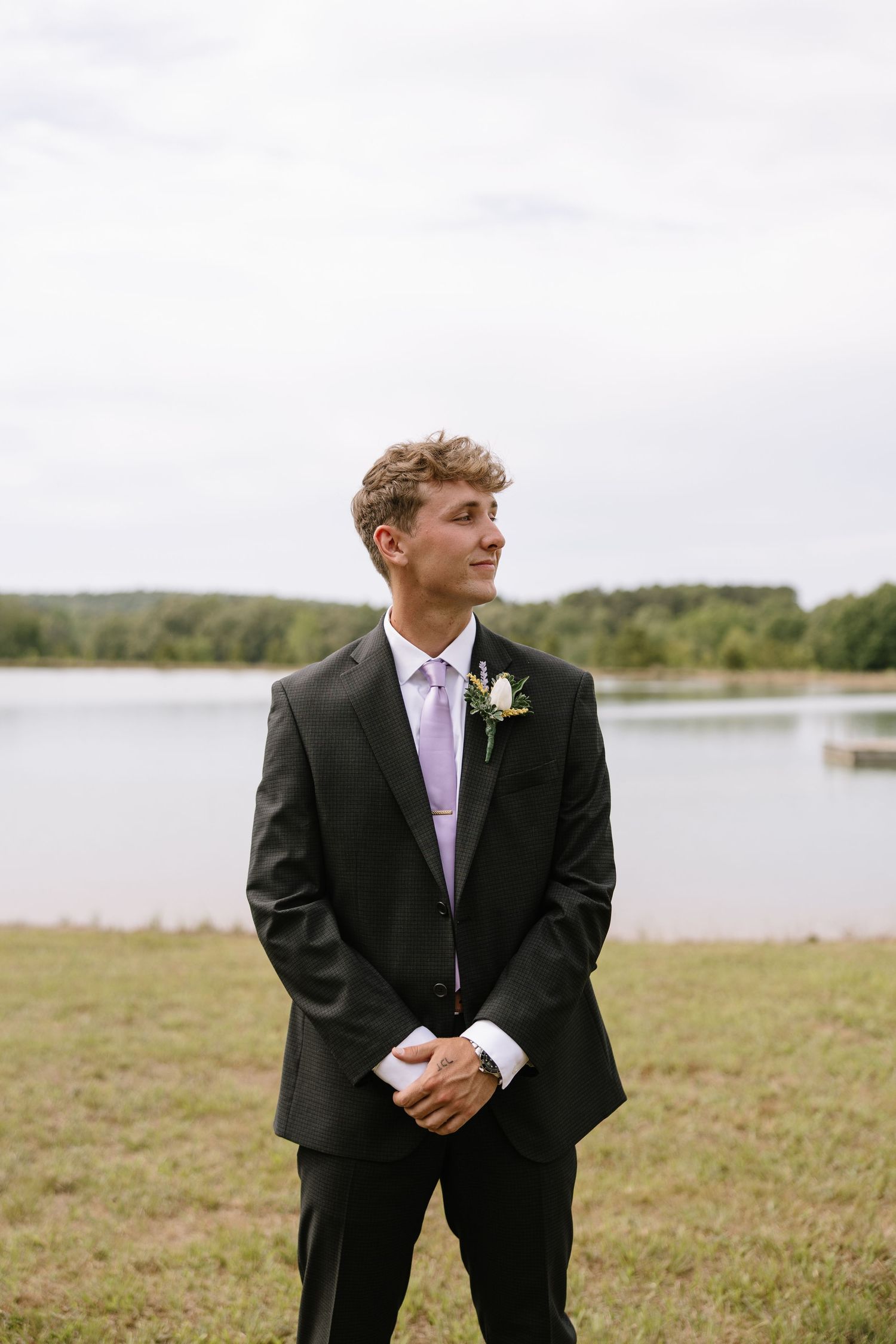 Young groom in black suit with lavender tie and boutonniere stands by lakeside on overcast wedding day.