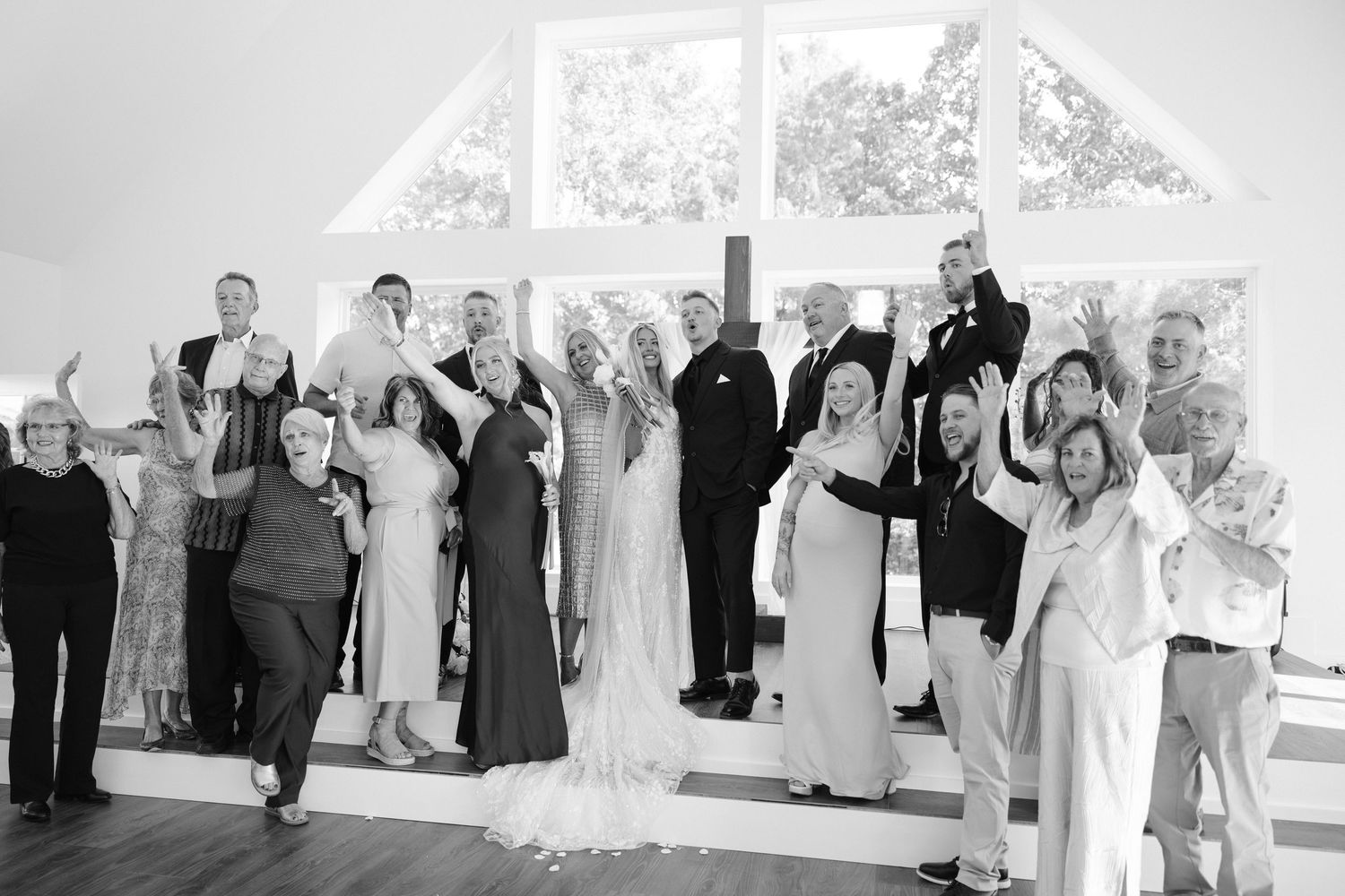 Black and white wedding photo of a large group celebrating joyfully inside a chapel with tall windows.