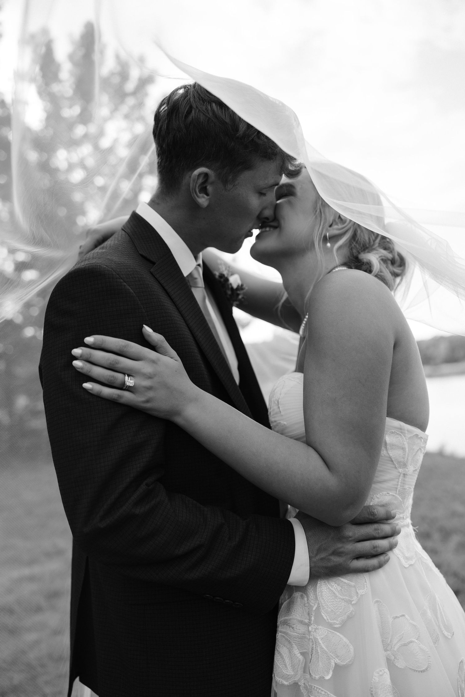 Black and white romantic photo of bride and groom nearly kissing under flowing veil outdoors.