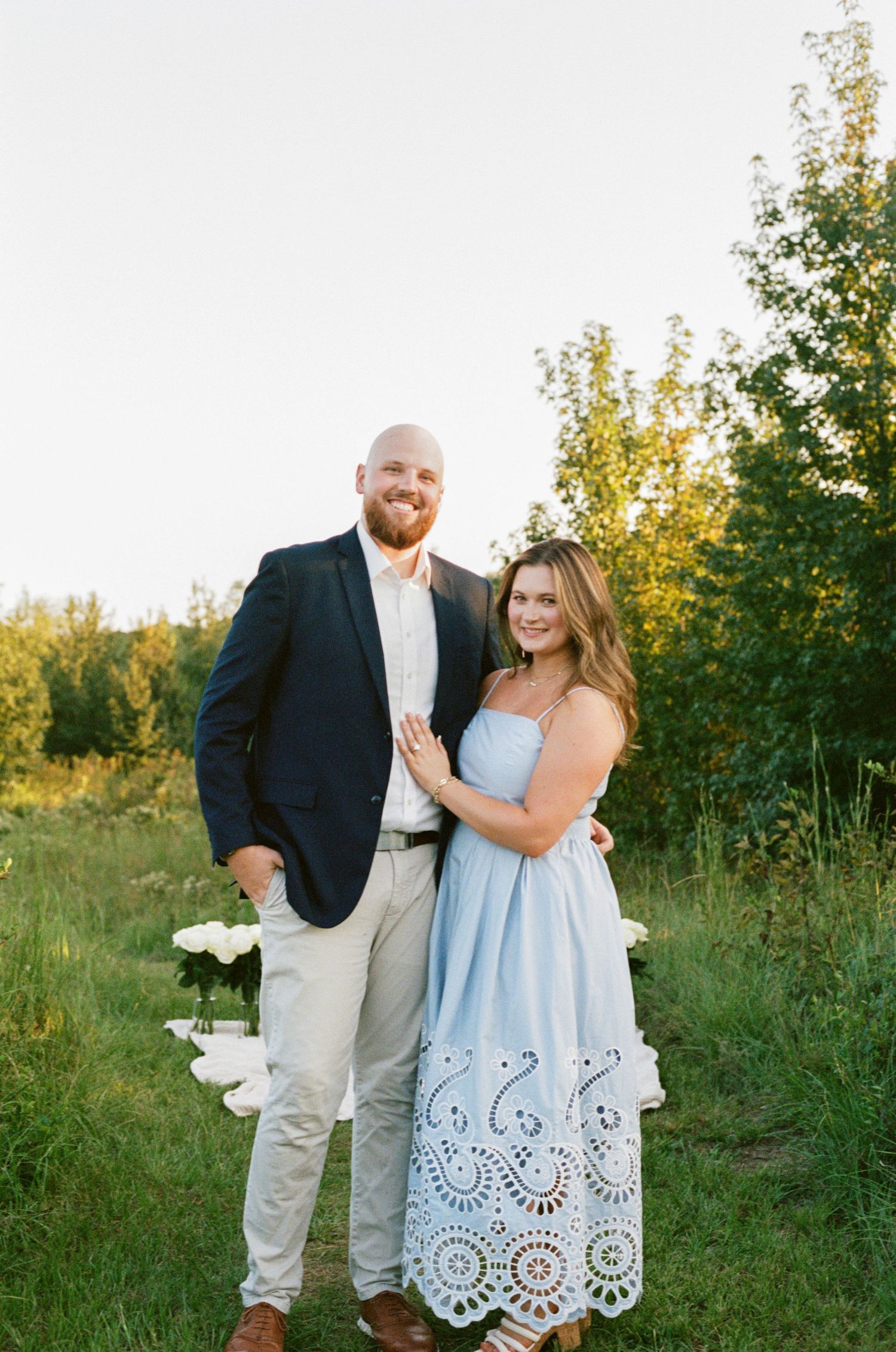 Couple posing outdoors in golden hour light, man in navy blazer, woman in light blue eyelet dress, surrounded by greenery.