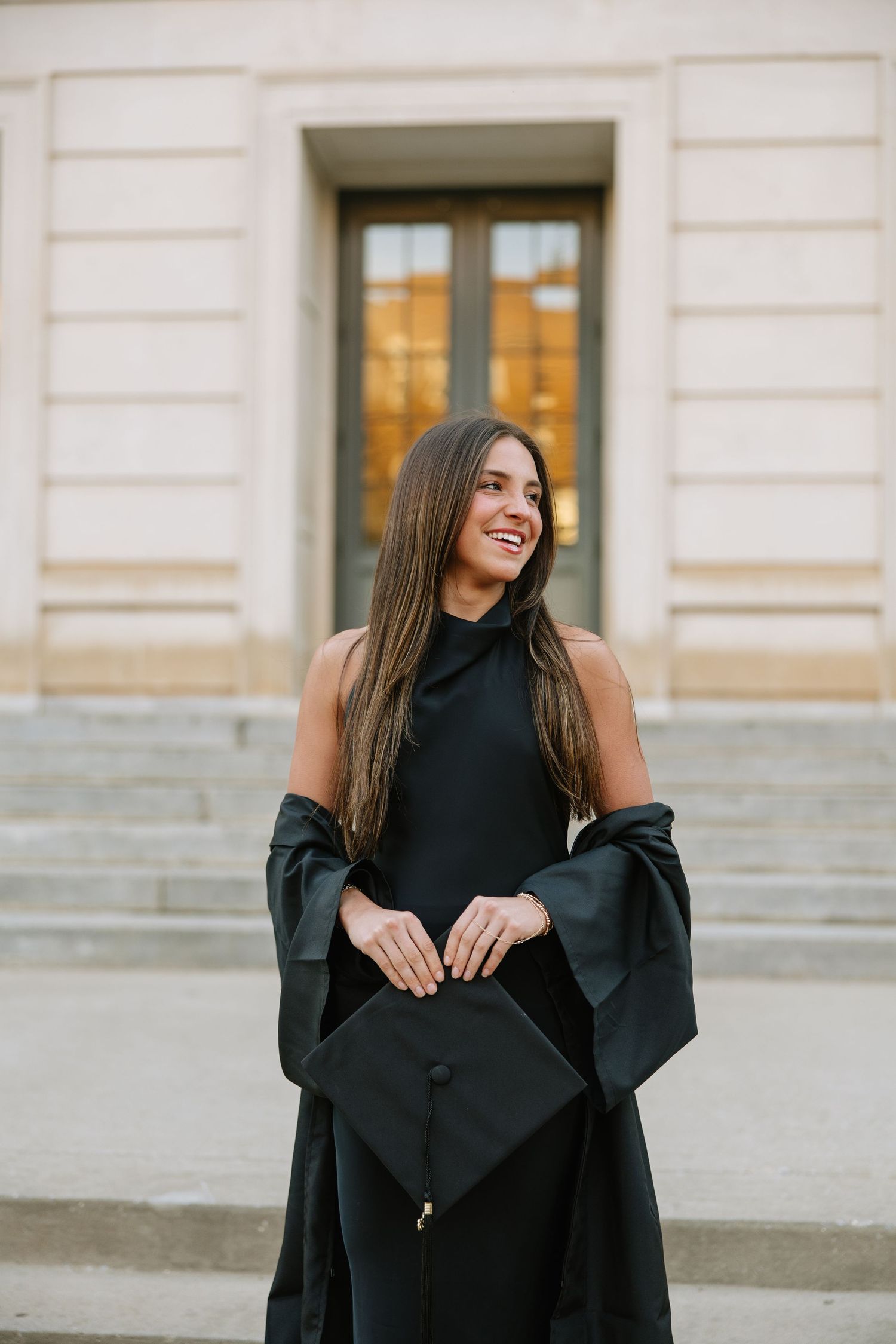 Smiling young woman in black halter dress holding graduation cap on university building steps.