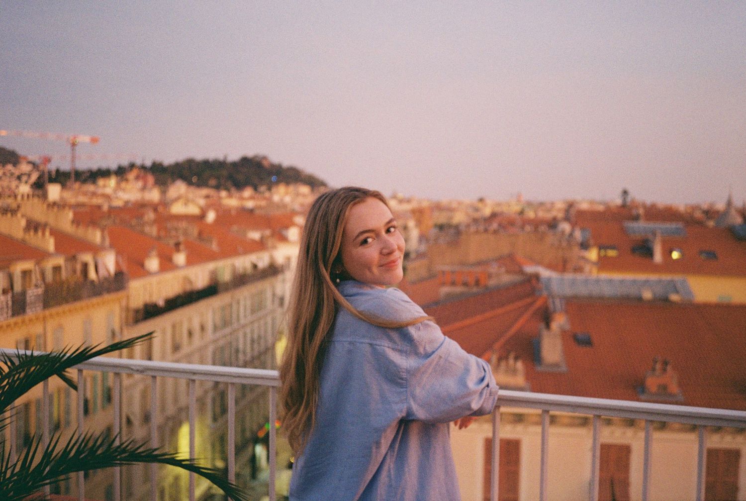 Young blonde woman smiling over shoulder on balcony overlooking European city rooftops at sunset.