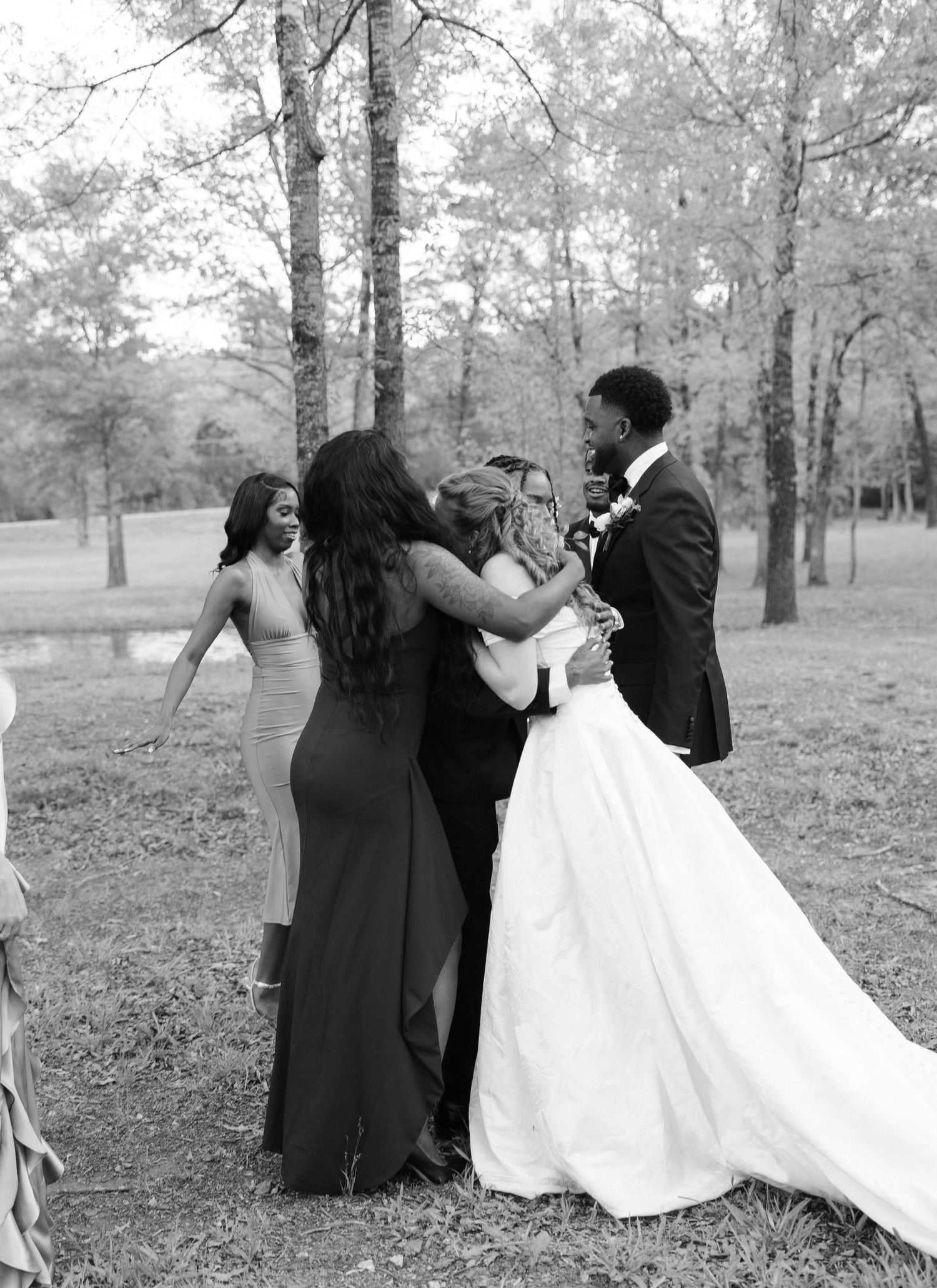 Black and white photo of wedding party congratulating the interracial bride and groom outdoors among trees.