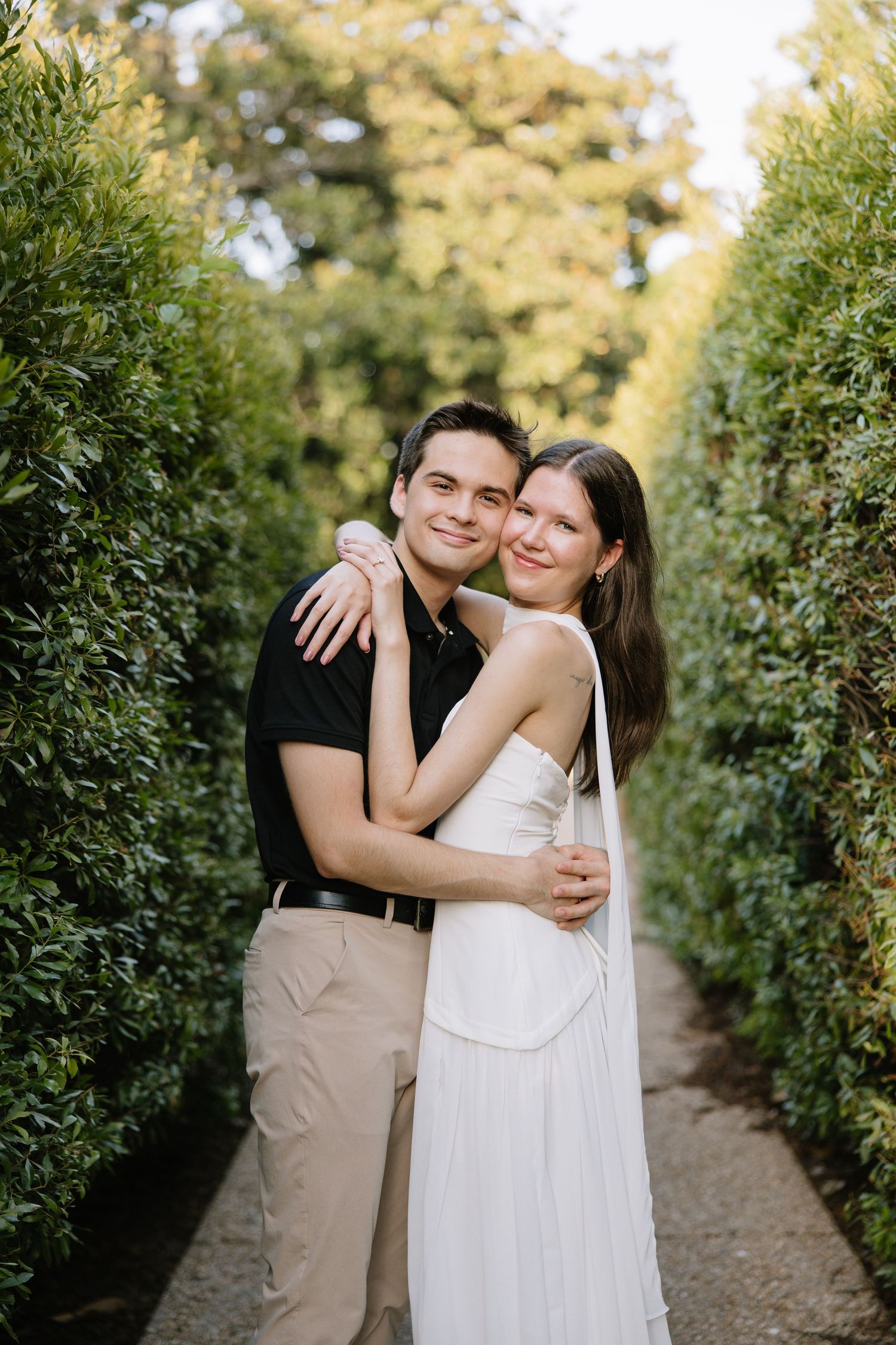 Smiling couple hugging between tall green hedges in a garden pathway.