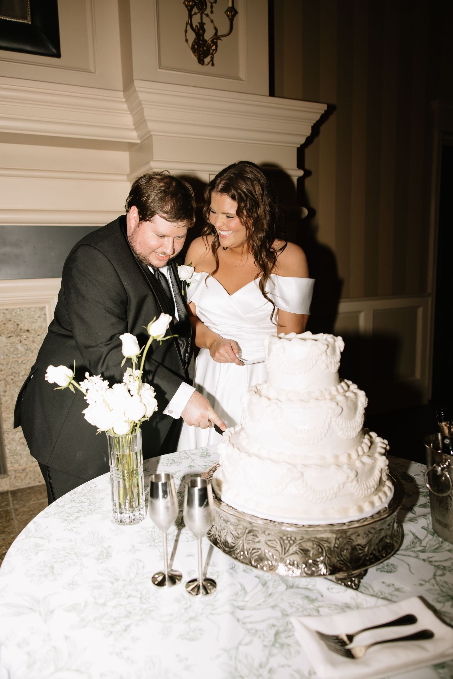 Bride and groom cut tiered white wedding cake together at elegant reception with champagne flutes nearby.