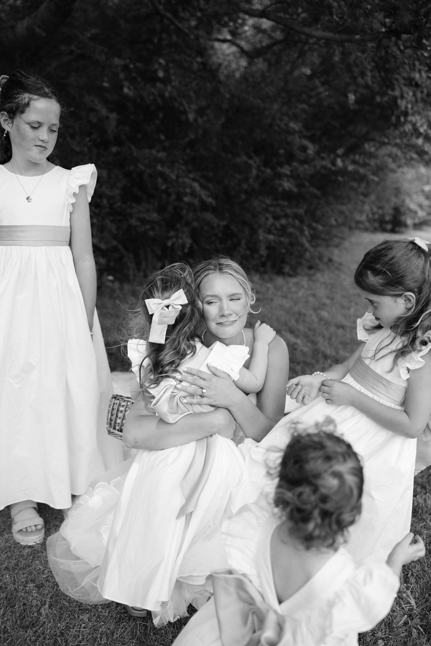Black and white photo of bride kneeling and hugging flower girls in white dresses outdoors.