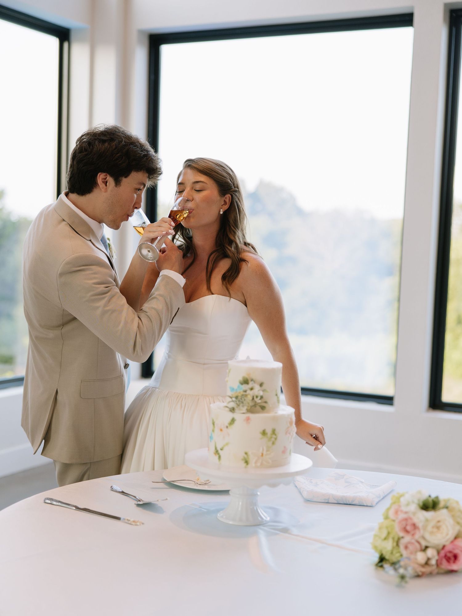 Bride and groom sharing cake and champagne at wedding reception table with floral bouquet and large windows backdrop.