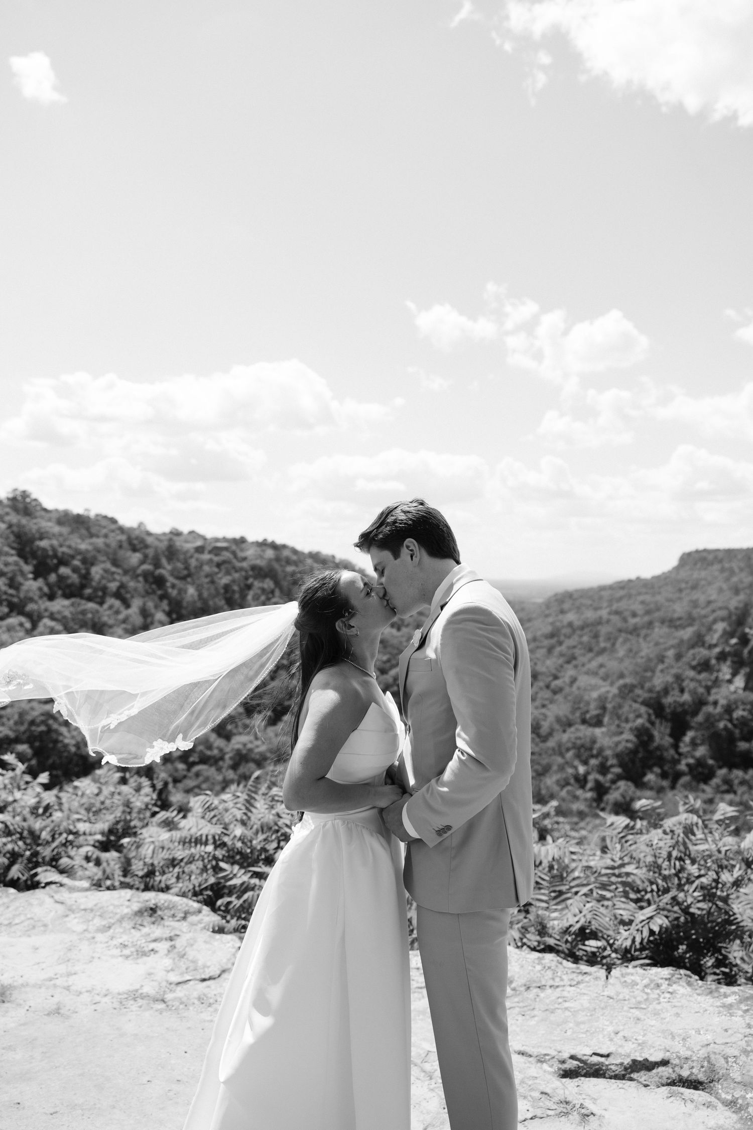 Black and white photo of bride and groom embracing on a rocky overlook, veil blowing in wind with forest backdrop.