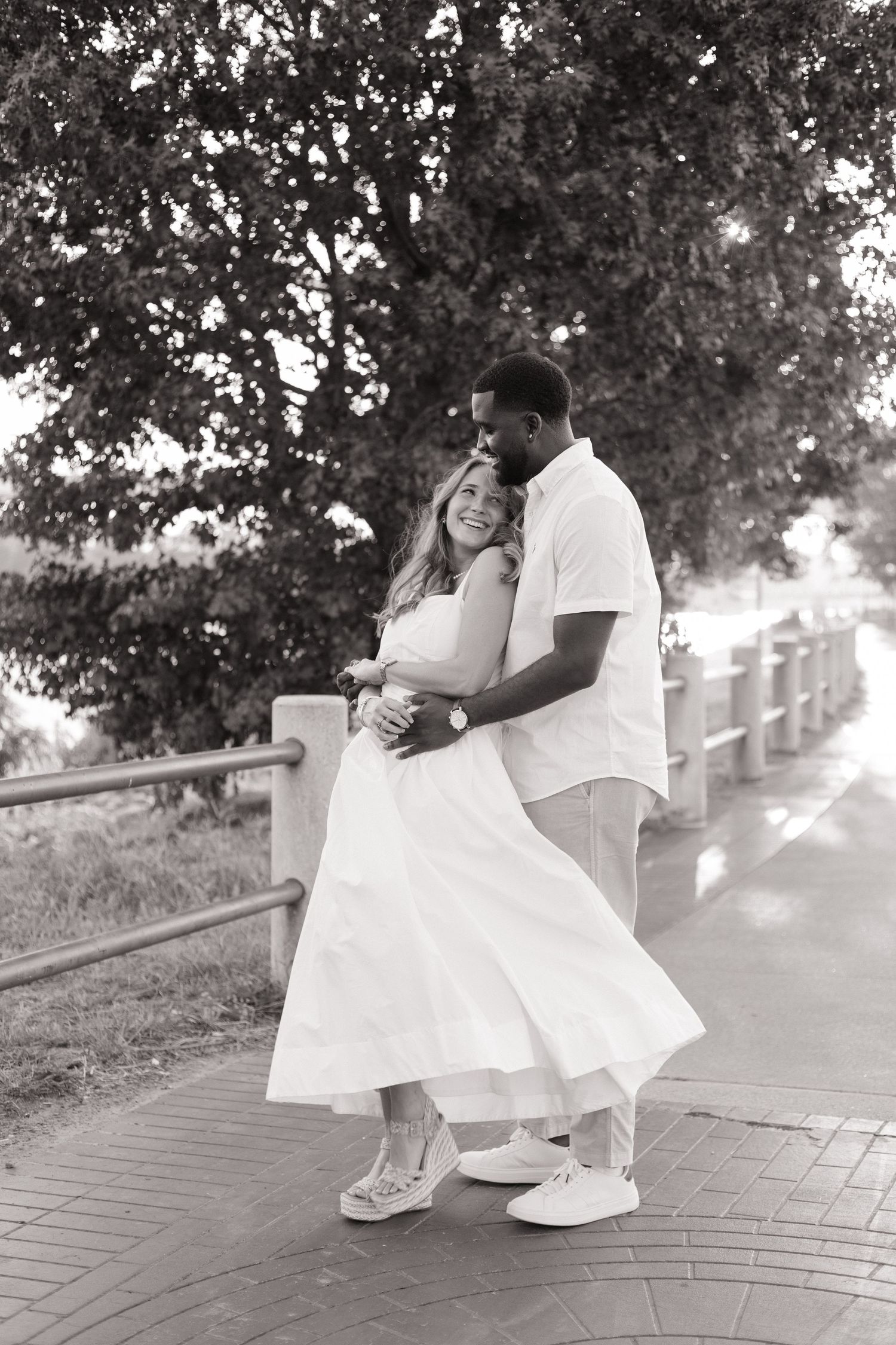 Black and white photo of a smiling couple embracing on a brick walkway, surrounded by lush trees.