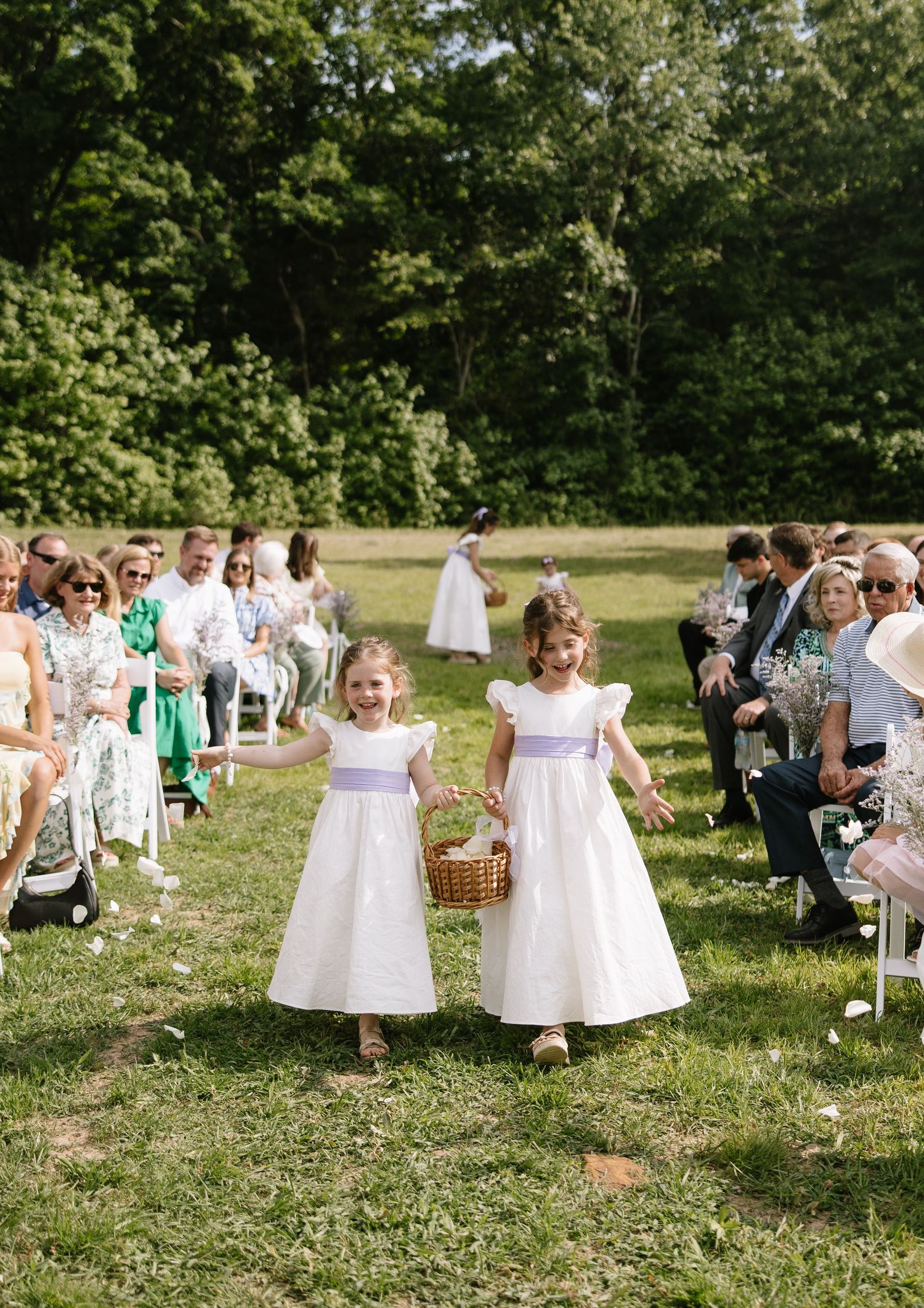 Two flower girls in white dresses with lavender sashes walk down an outdoor wedding aisle carrying a basket.