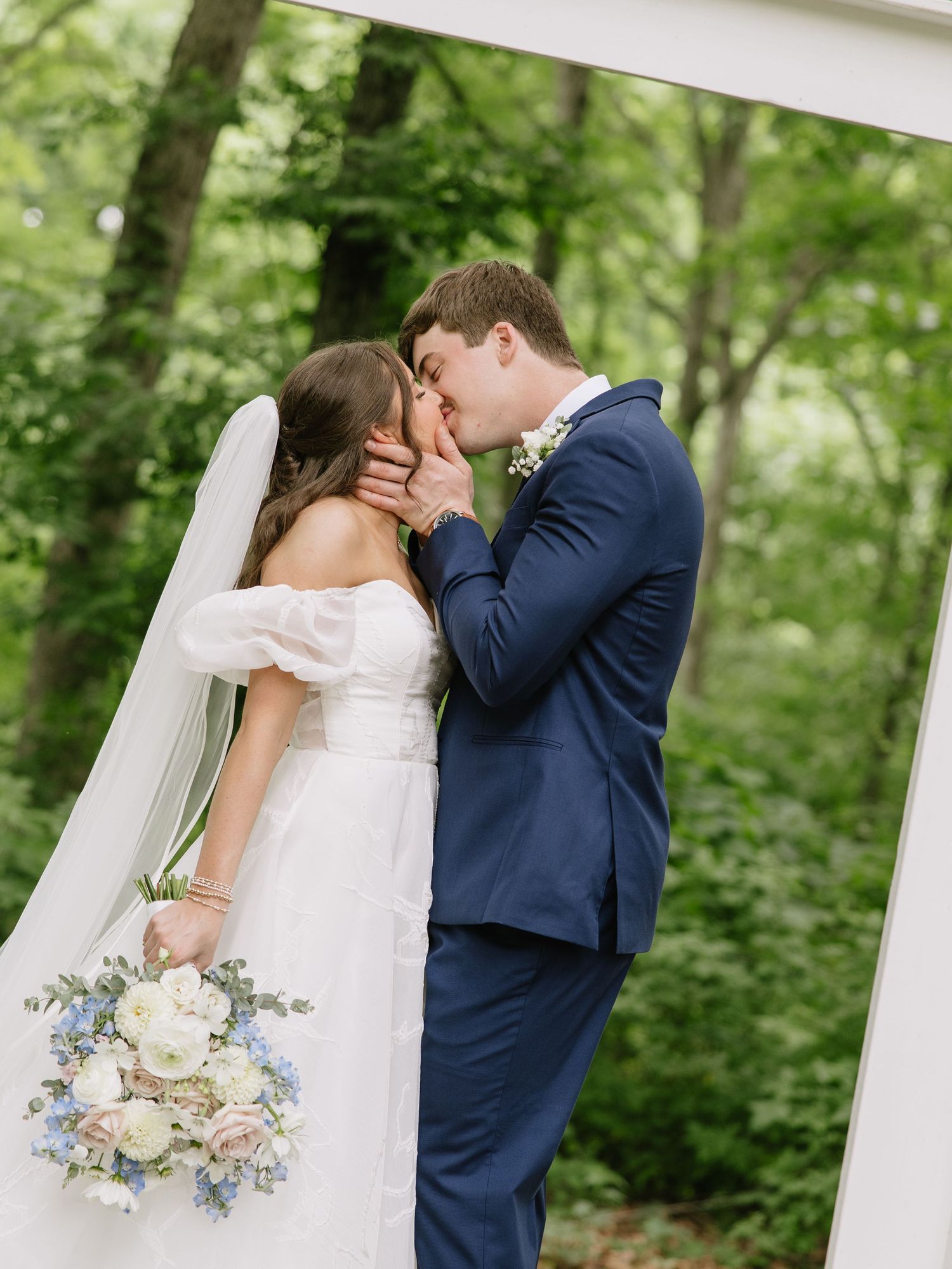 Bride and groom share a kiss outdoors, bride holding blue and white bouquet, groom in navy suit, surrounded by lush greenery.