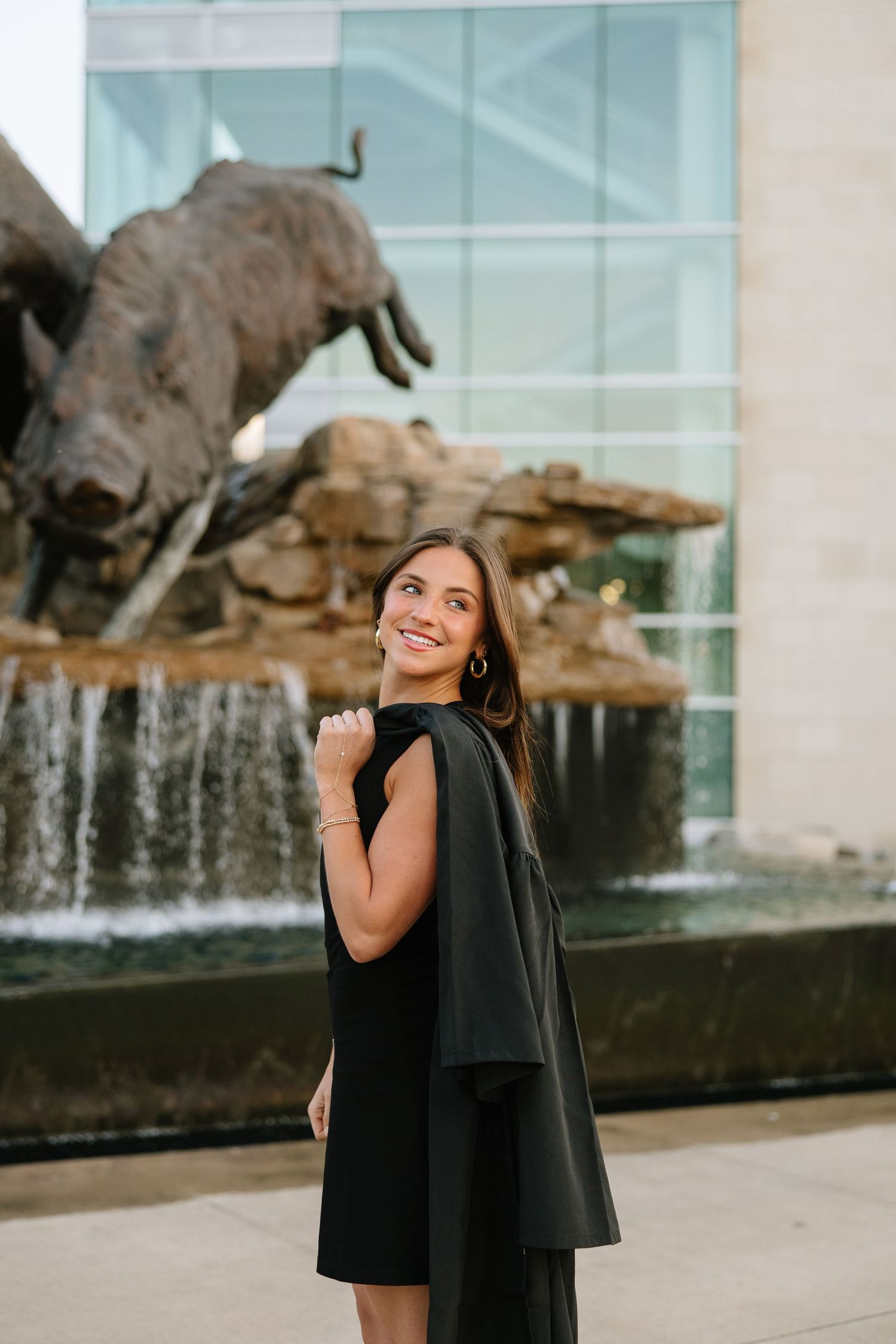 Smiling graduate in black dress and gown posing in front of the Razorbacks bronze boar fountain sculpture.
