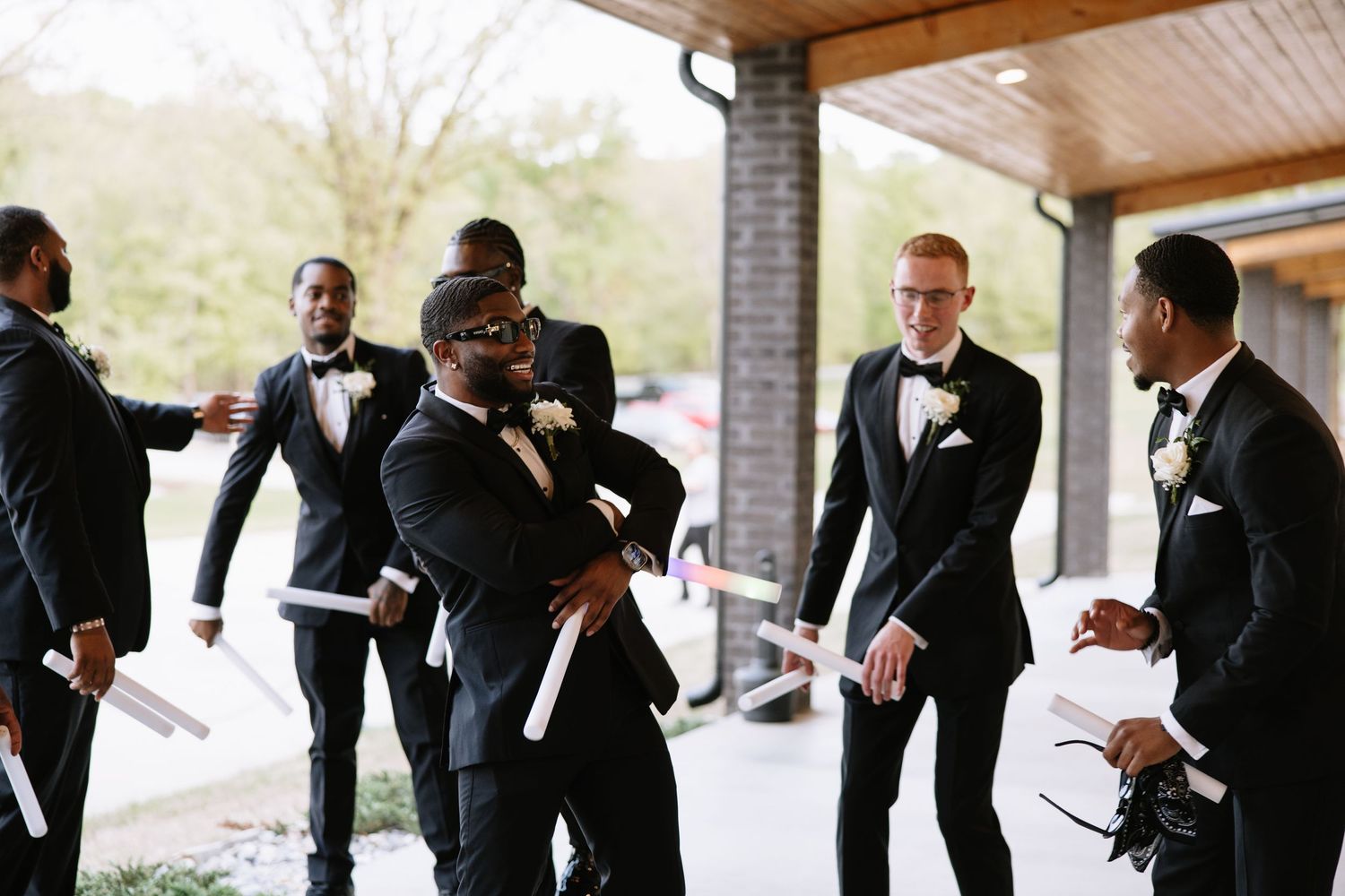 Groomsmen in black tuxedos laughing and holding programs outside wedding venue.