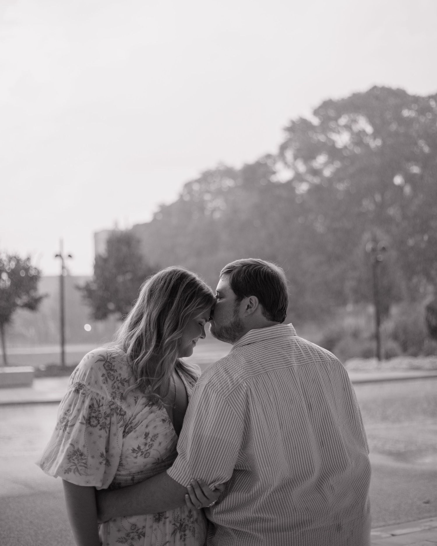 Black and white photo of a romantic couple leaning foreheads together outdoors near trees.