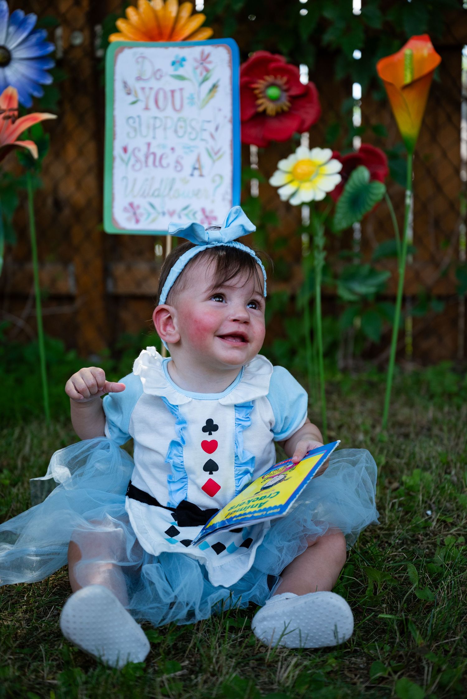 Baby dressed as Alice in Wonderland sits on grass with giant flowers and a quote sign backdrop.