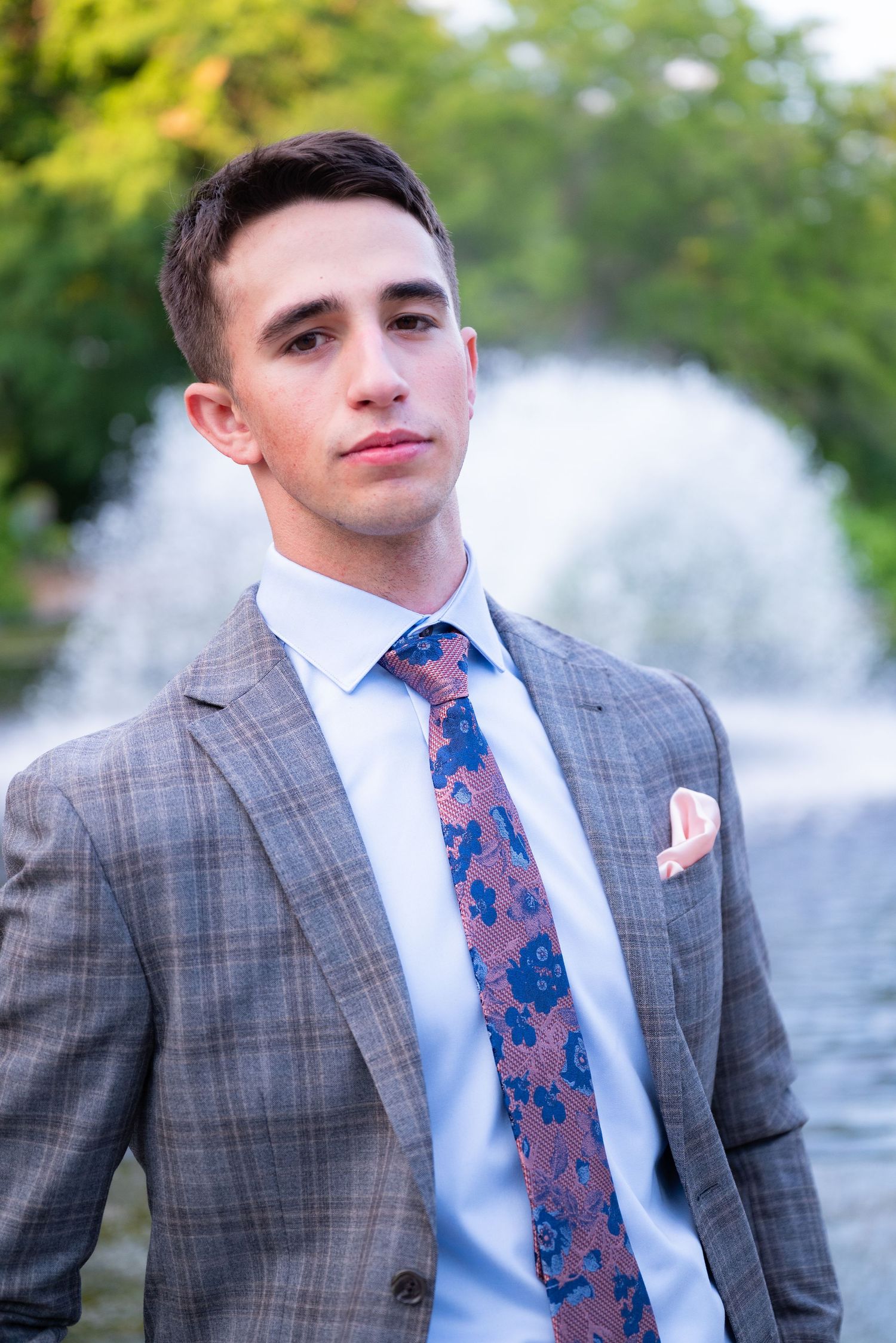 Young man in plaid suit with floral tie and pocket square poses near fountain outdoors.