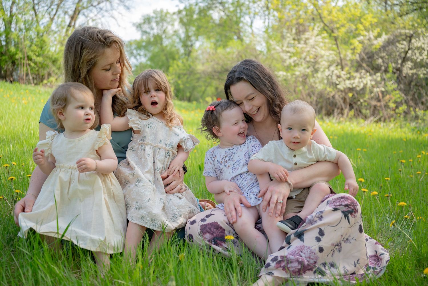 Two mothers sit in green grass with four young children on a sunny spring day outdoors.