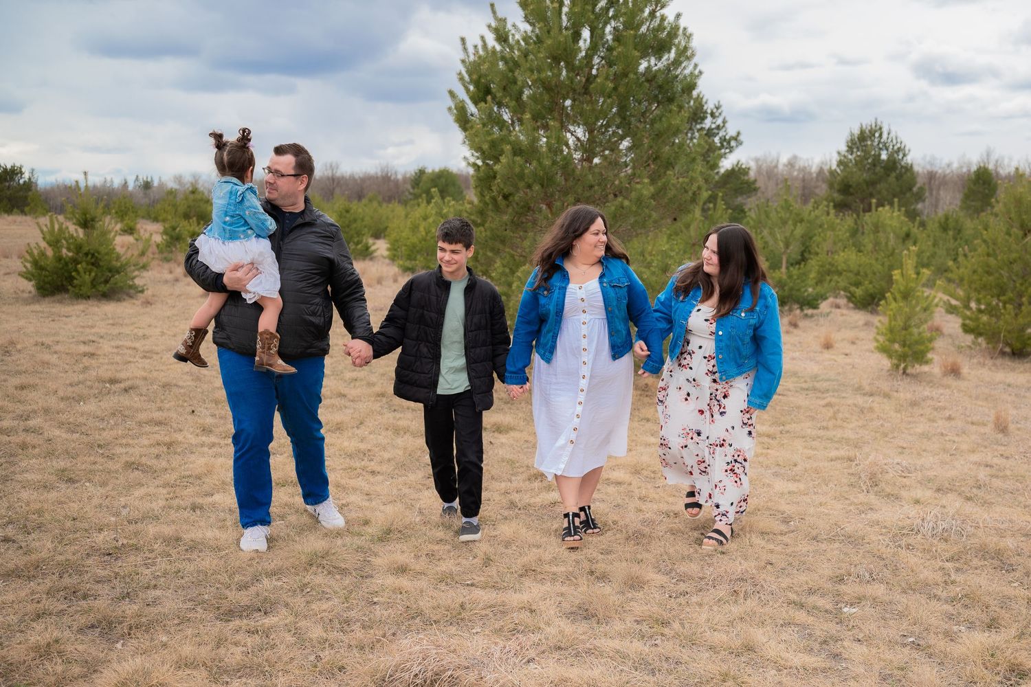 Family of five walking hand-in-hand through a pine tree field on a cloudy day, smiling and enjoying outdoor time.