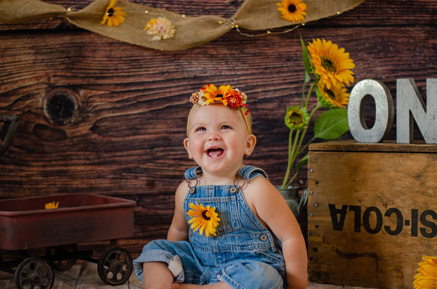 Happy baby in denim overalls with floral headband laughing in a rustic sunflower-themed photo session.