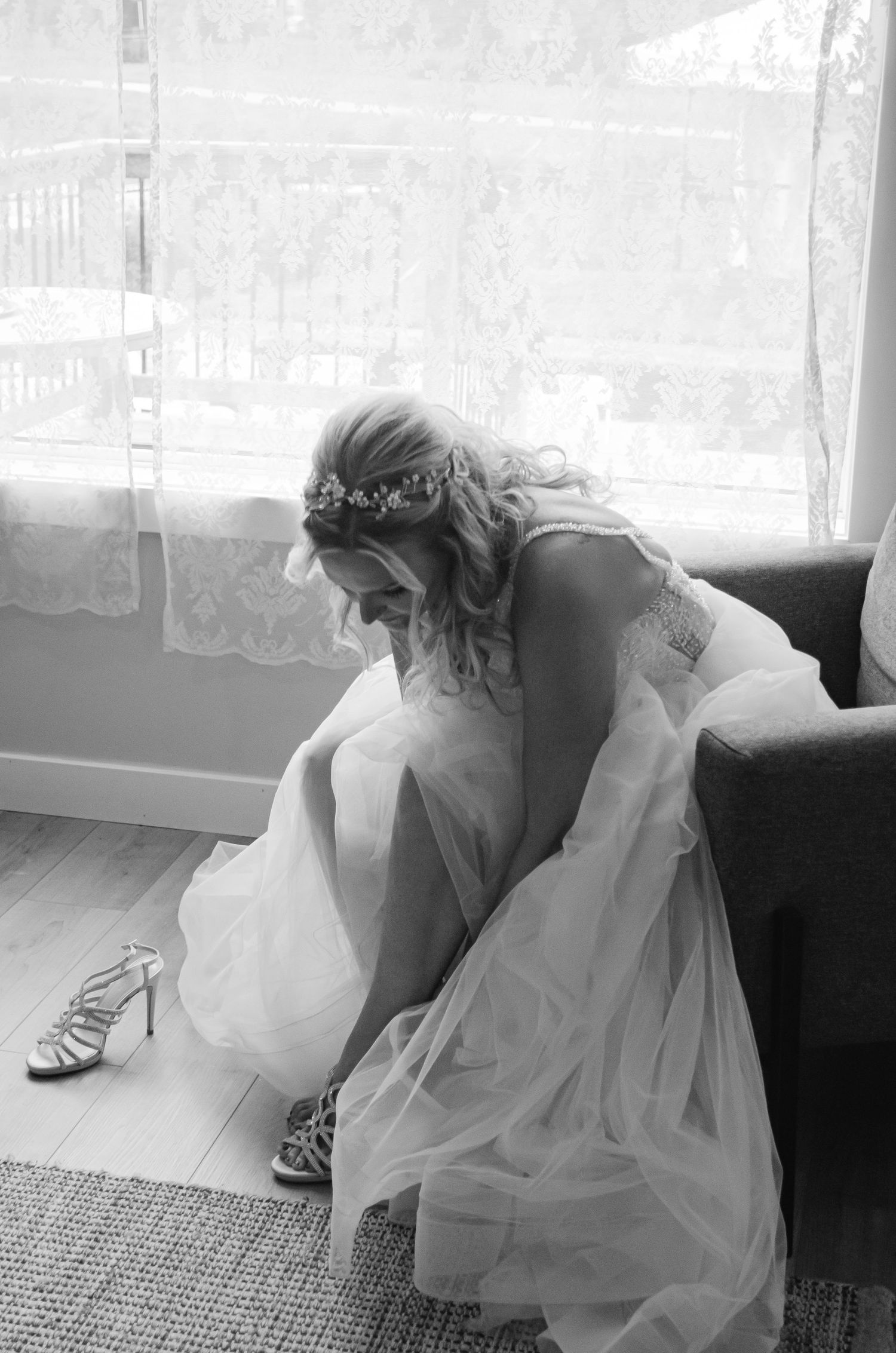 Black and white photo of a bride in a white gown and floral crown putting on heeled sandals by a window.