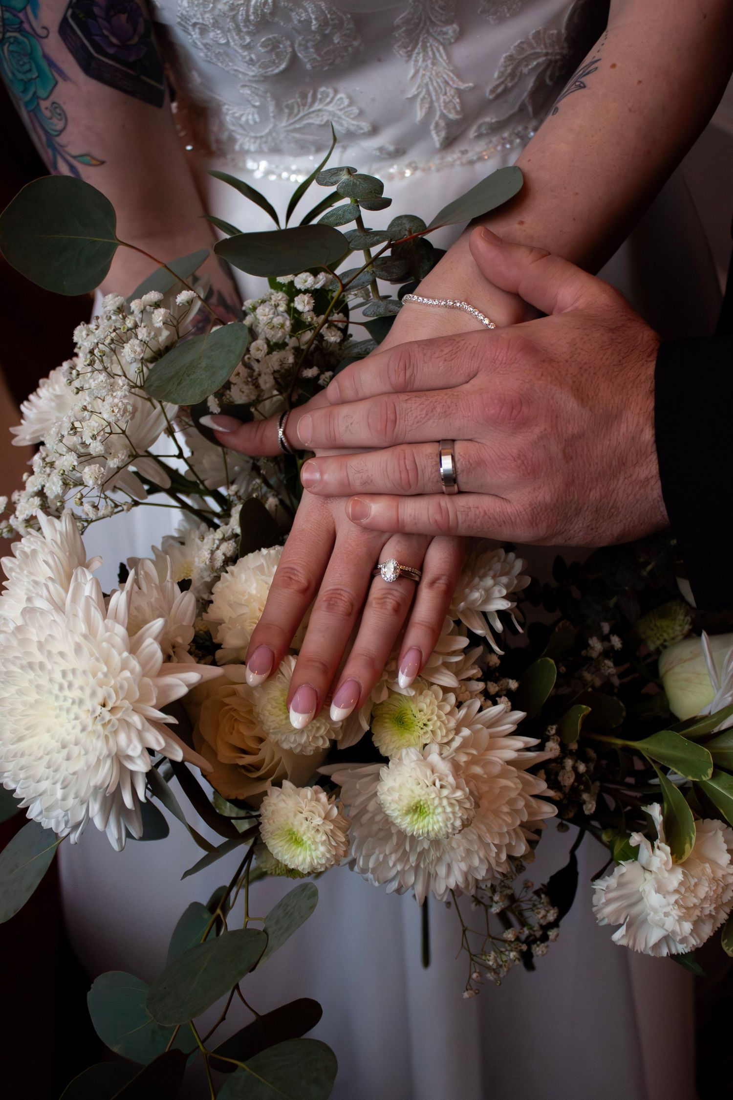 Bride and groom display wedding rings over a bouquet of white chrysanthemums and eucalyptus.