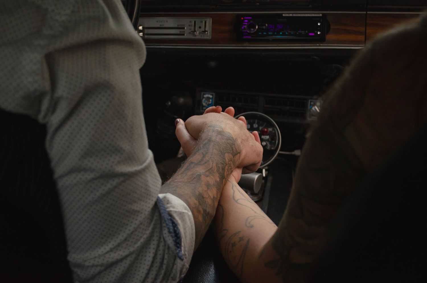 Two people holding hands over a car gear shift, one with a tattoo, inside a vintage vehicle.