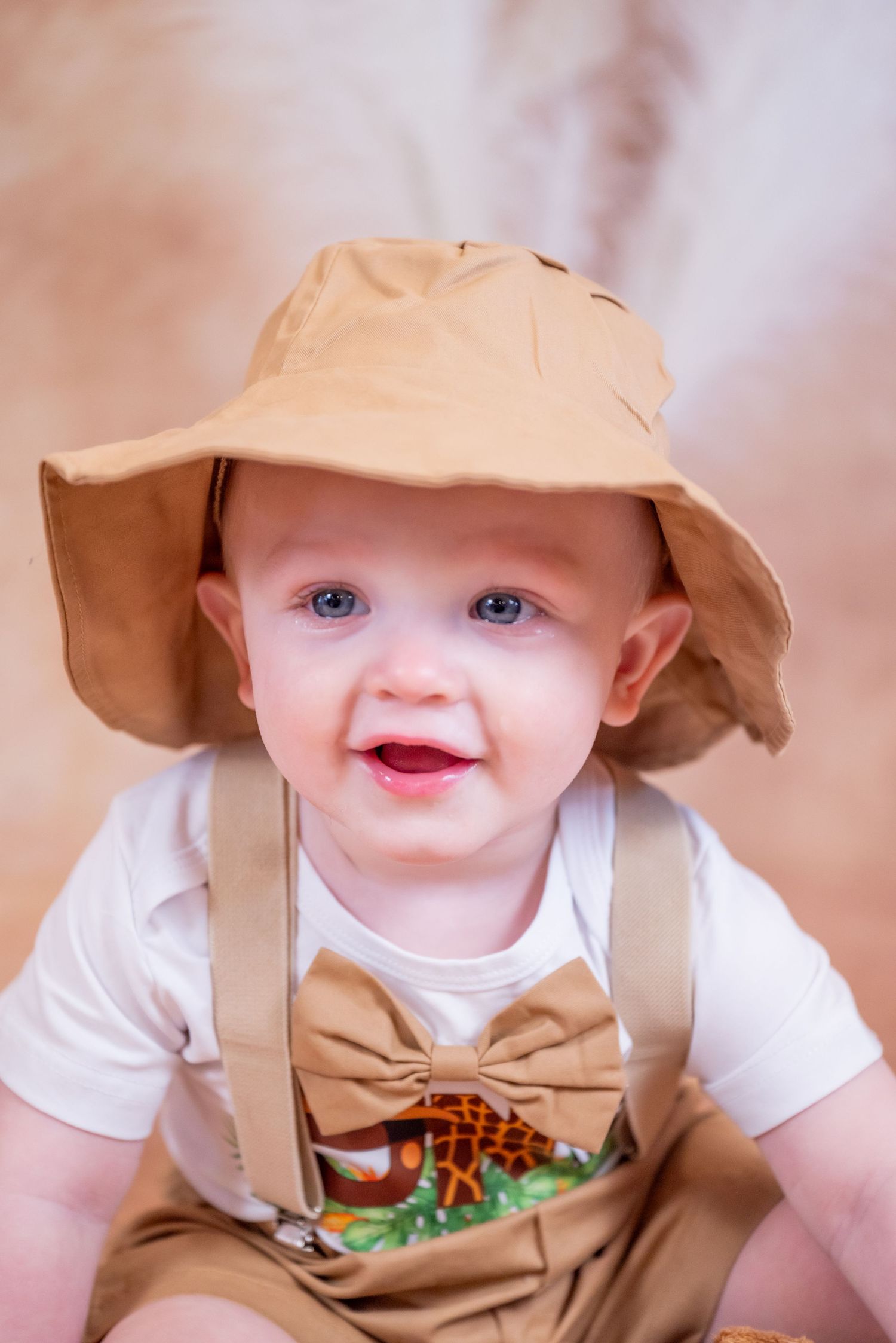 Adorable baby boy smiling in tan safari hat, bow tie suspenders, and white onesie with giraffe print.