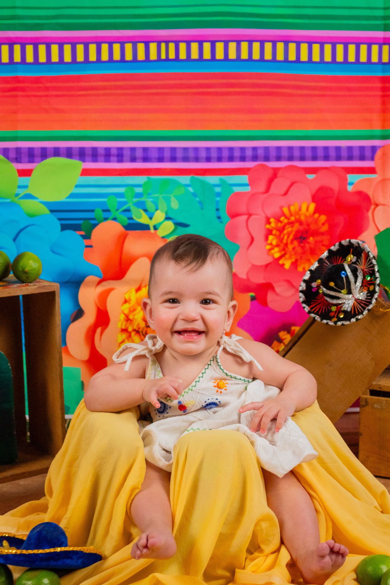 Happy baby in yellow dress sits before colorful Mexican fiesta backdrop with paper flowers and sombrero.