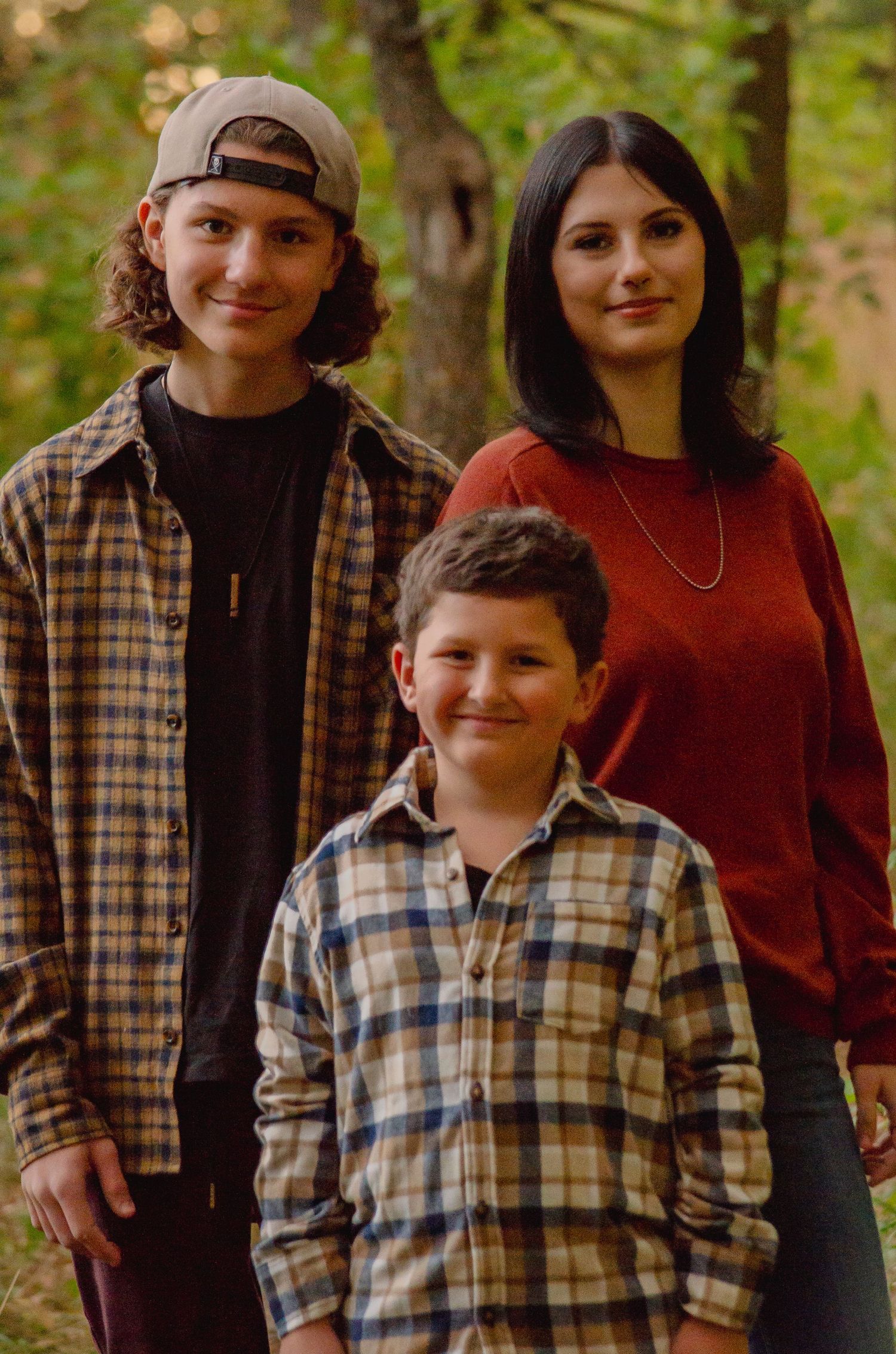 Two boys and a teenage girl pose together outdoors among autumn trees in plaid shirts for a family portrait.