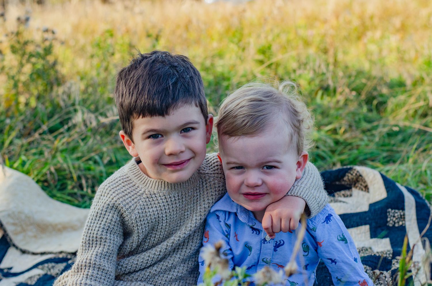 Two young brothers smiling and posing together on a blanket in a sunny field during a family outdoor photo session.