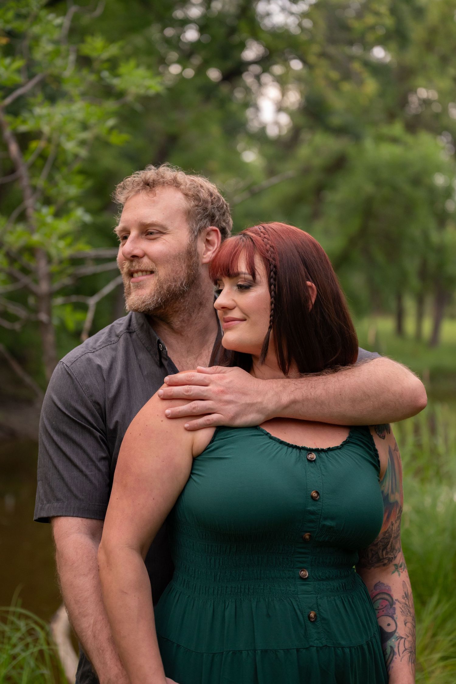 A couple embraces outdoors near a creek, woman in green dress with red hair, man in gray shirt, surrounded by lush trees.