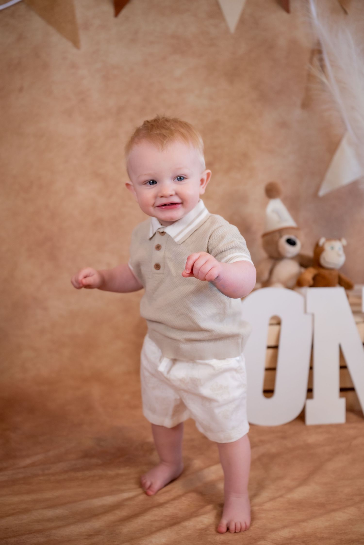 Smiling baby boy in beige outfit standing barefoot at a first birthday photo shoot with teddy bears.