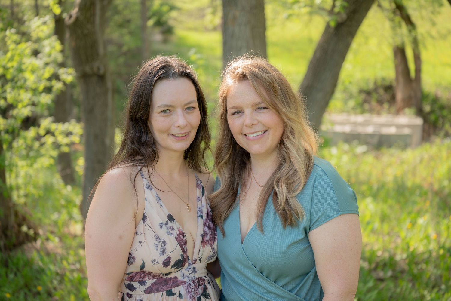 Two smiling women pose together outdoors among green trees in a sunny park setting.