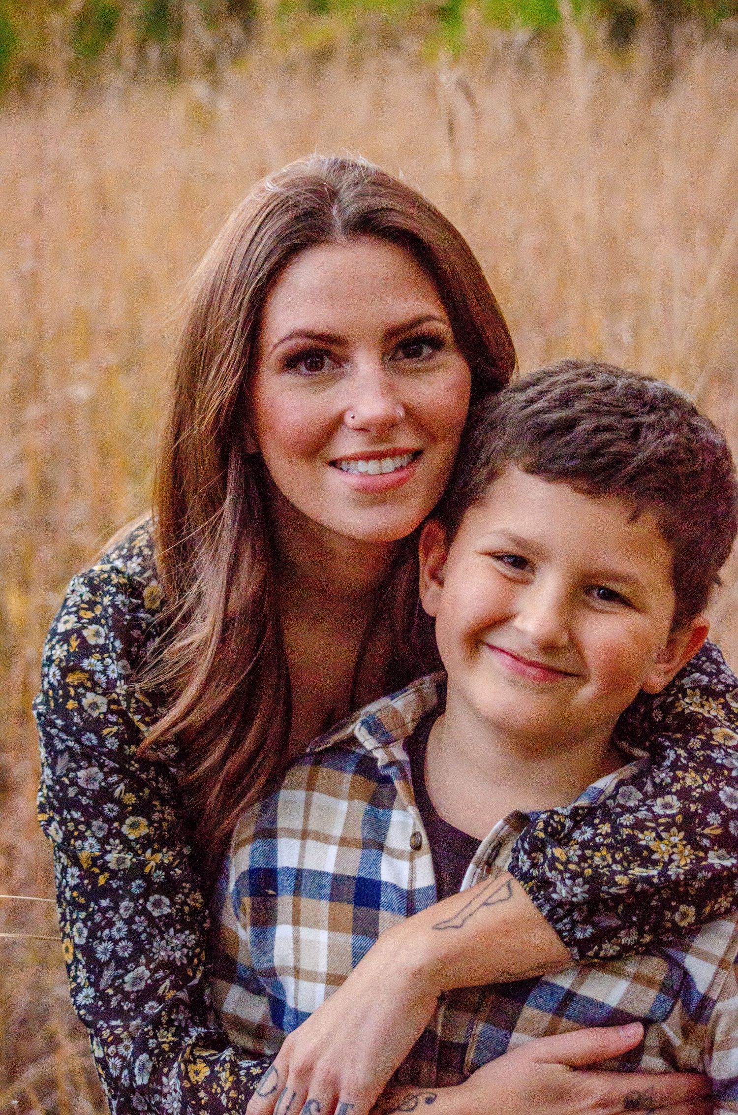 A smiling woman with brown hair hugs a young boy in a plaid shirt outdoors in a golden field.
