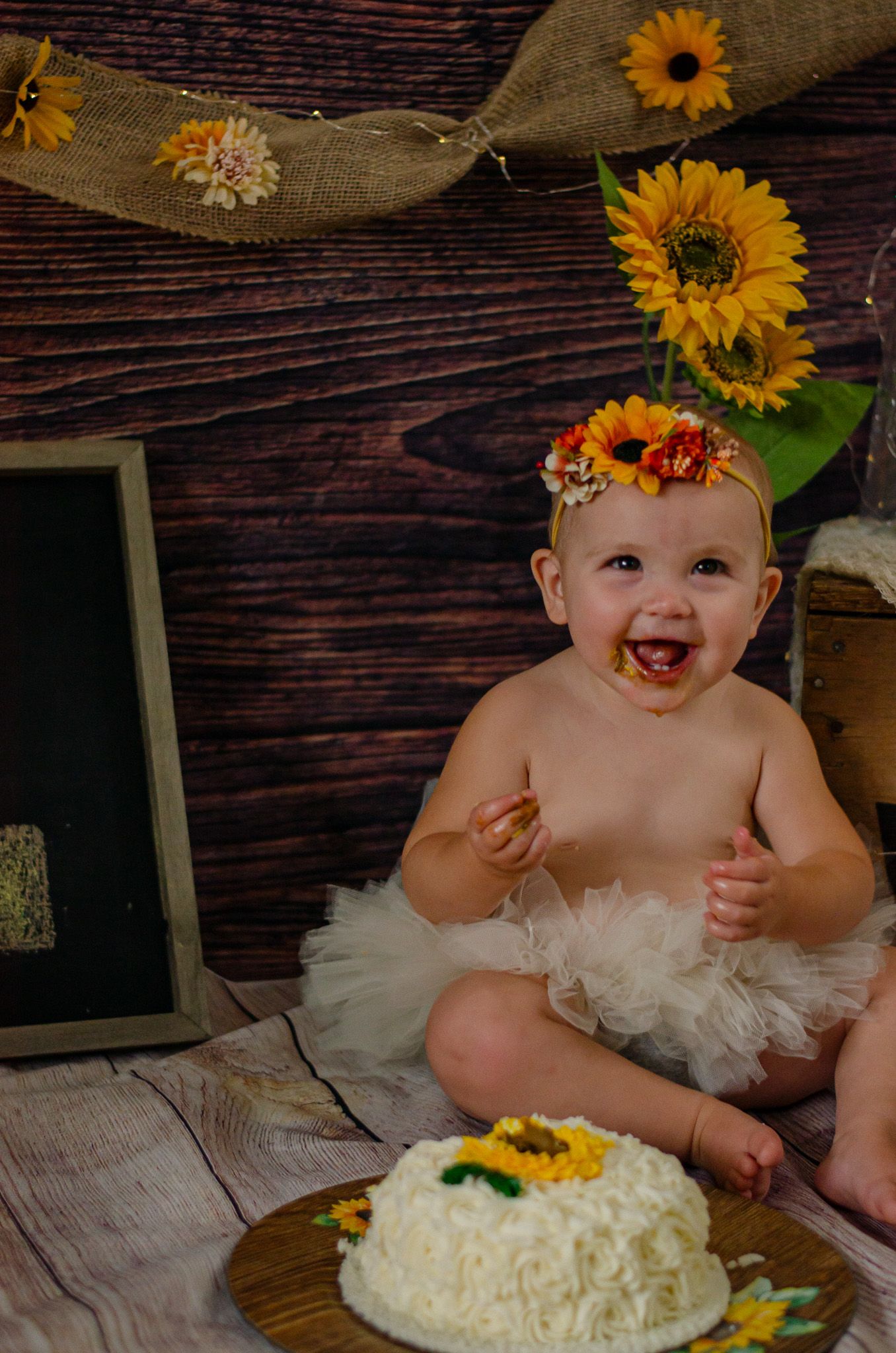 Baby girl in white tutu and sunflower crown smiles during first birthday cake smash with white sunflower cake.