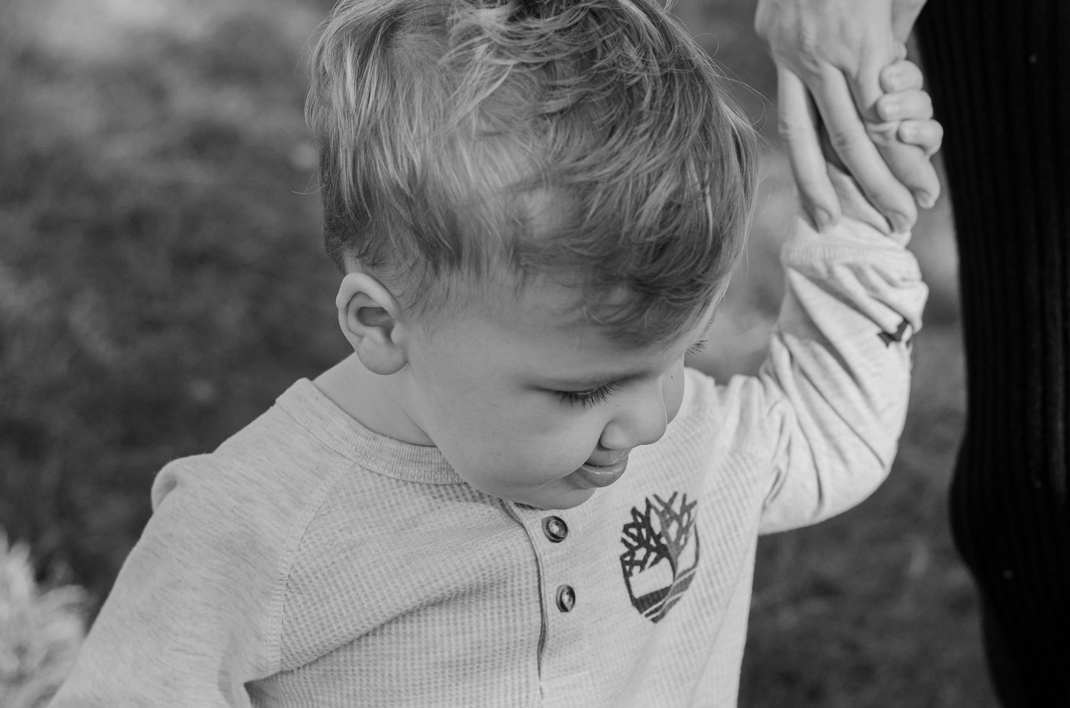 Black and white photo of a young toddler boy in a Timberland shirt holding an adults hand outdoors.