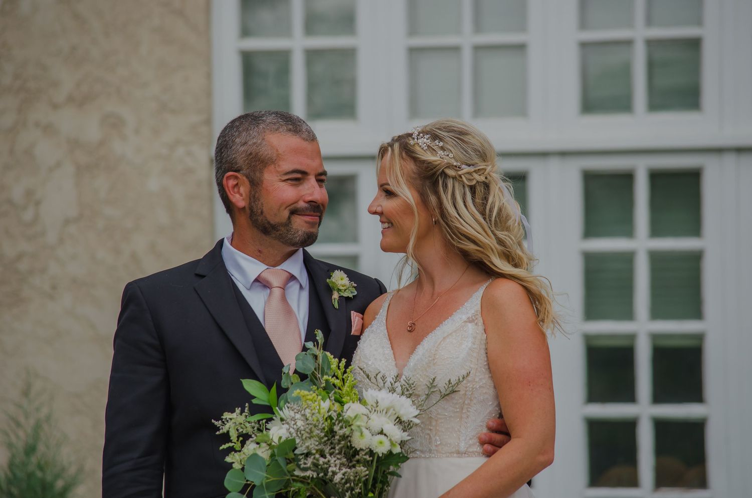 Smiling bride holding white bouquet and groom in navy suit with pink tie share a romantic moment outdoors.