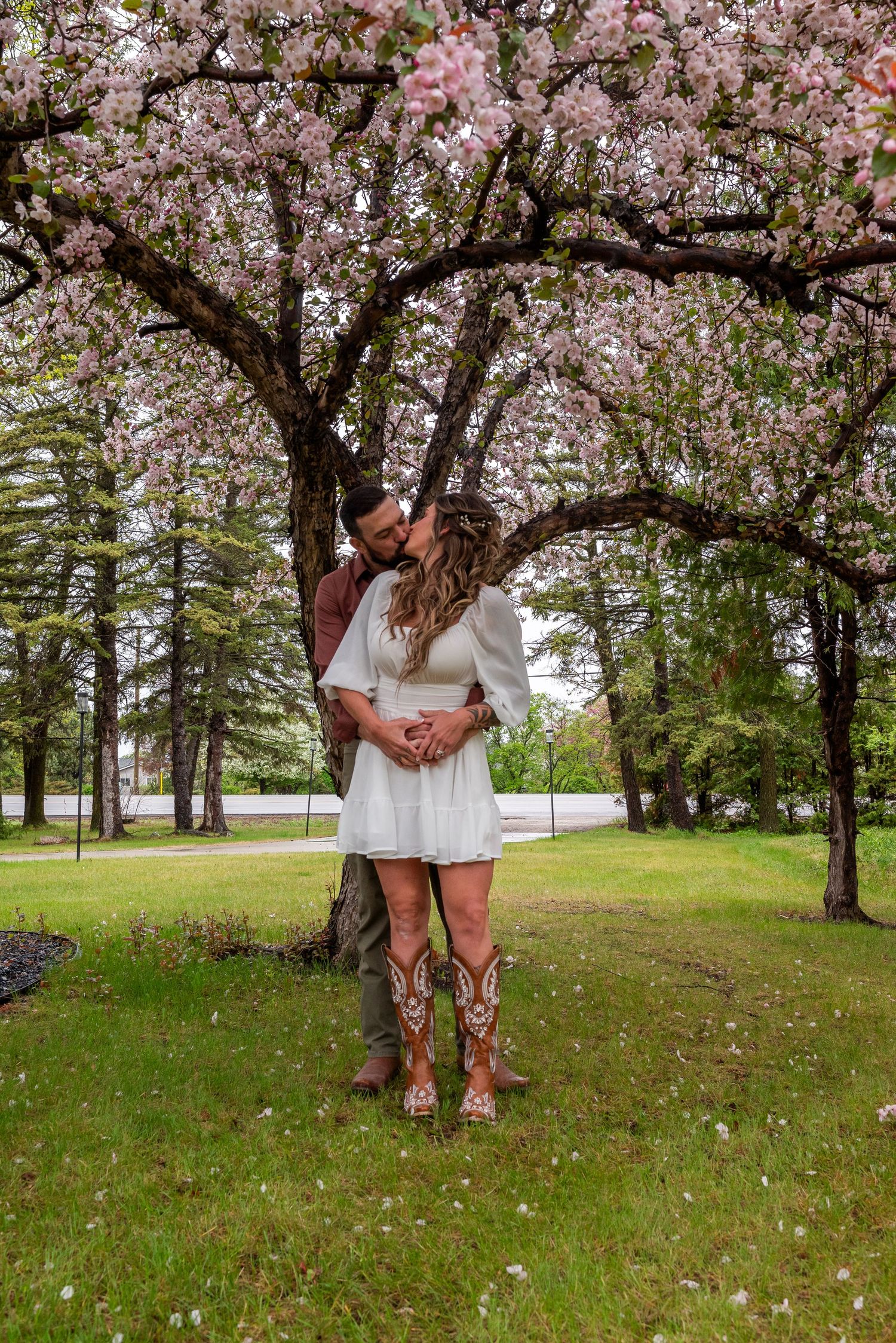 Couple kissing under a blooming pink cherry blossom tree on green grass, woman in white dress and cowboy boots.