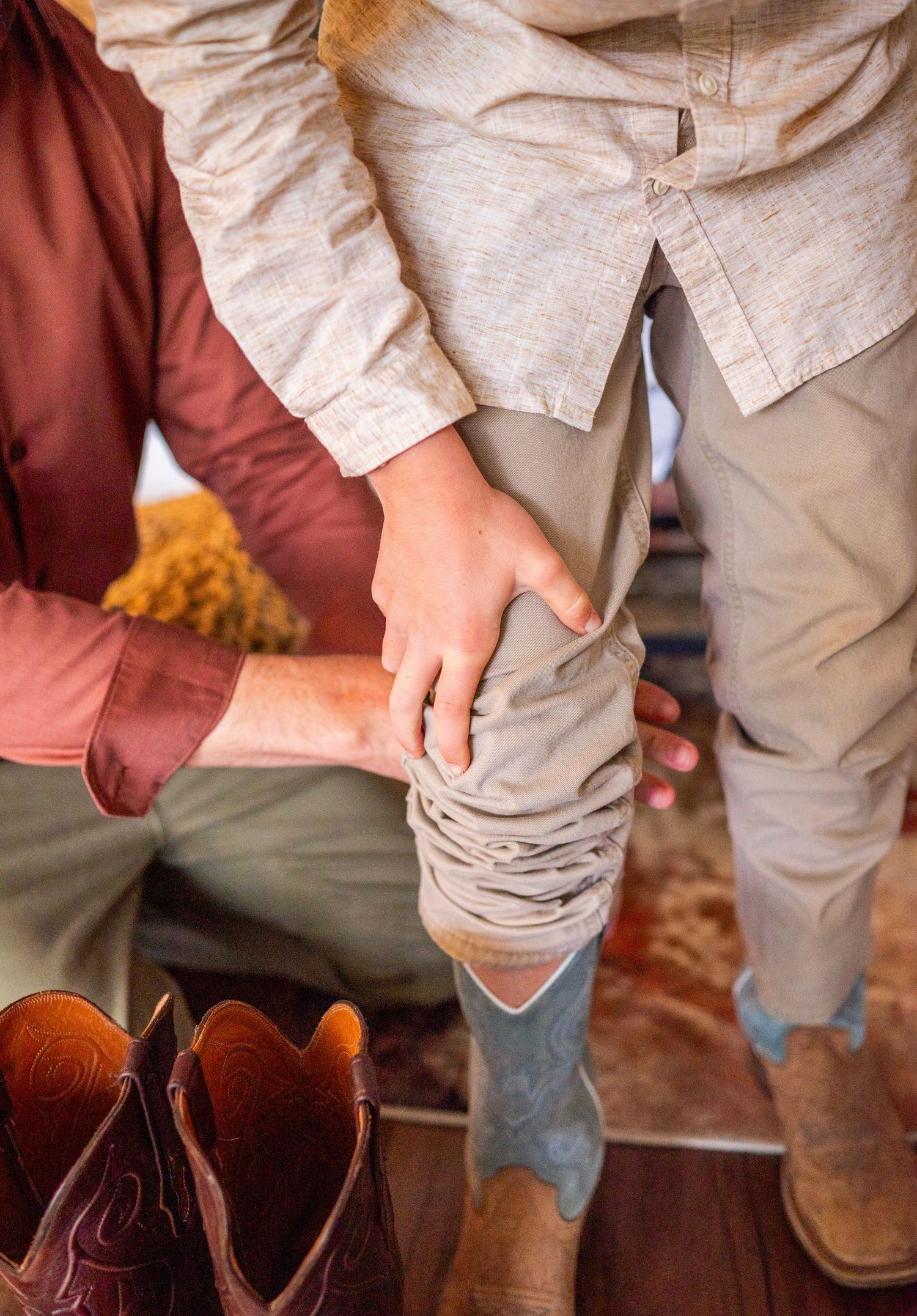 Adult helping a child put on brown cowboy boots while wearing a tan suit on a wooden floor.