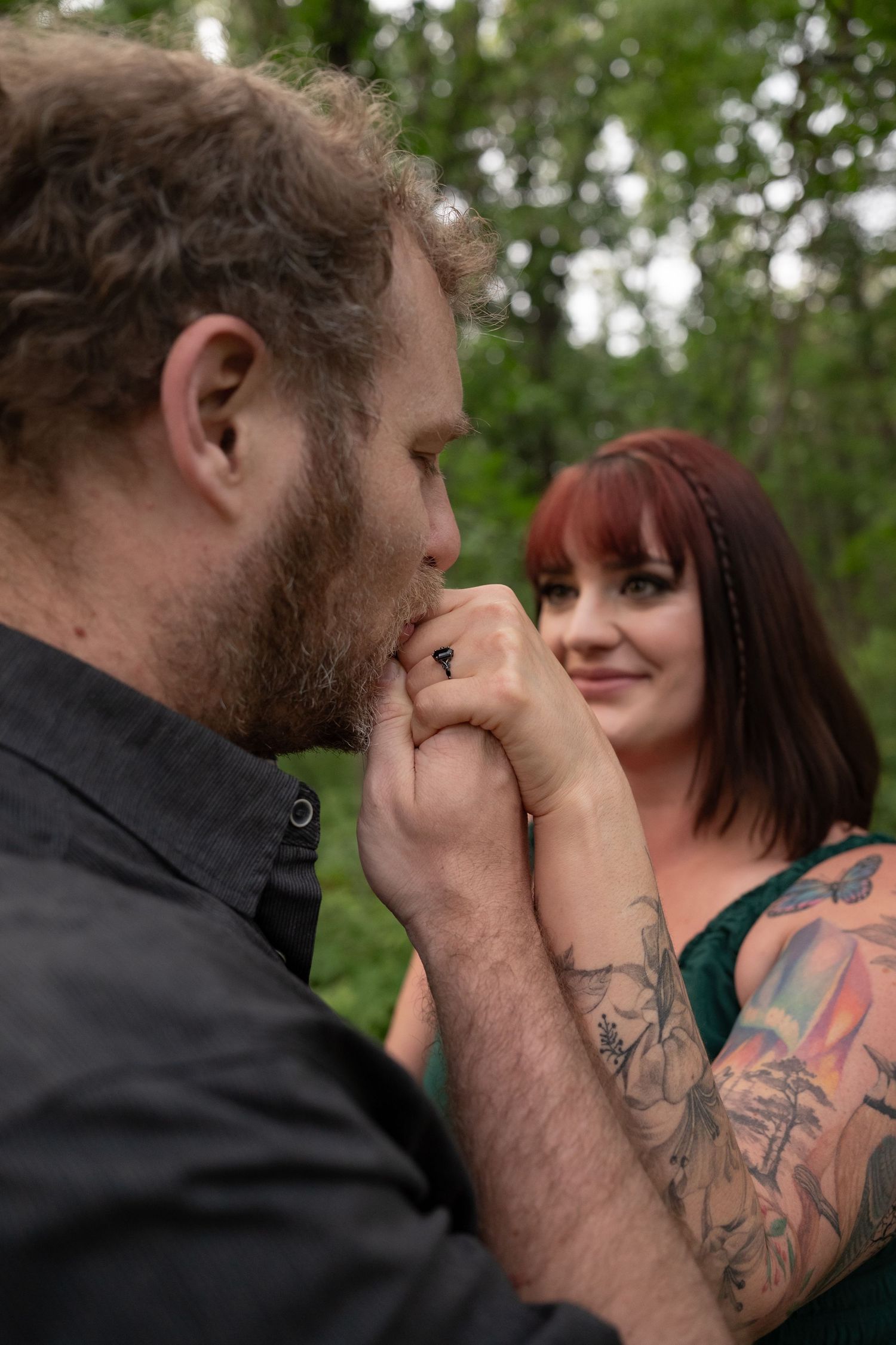 Man kissing tattooed woman's hand outdoors in a lush green forest setting.