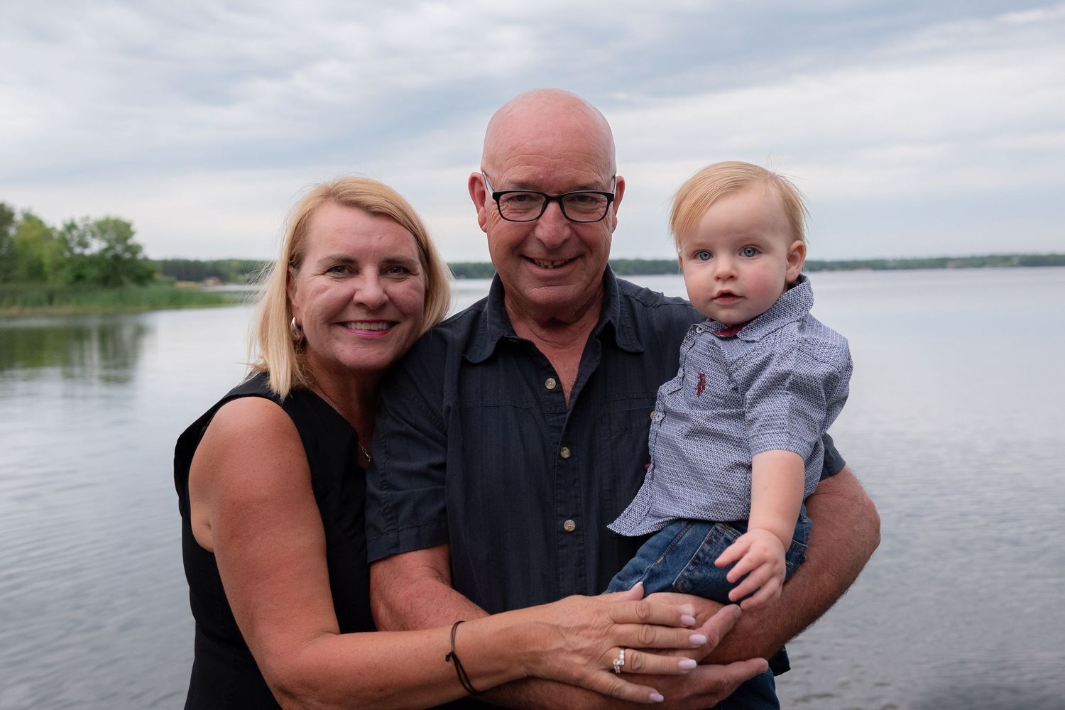 A smiling couple poses with a baby boy near a lake on a cloudy day.