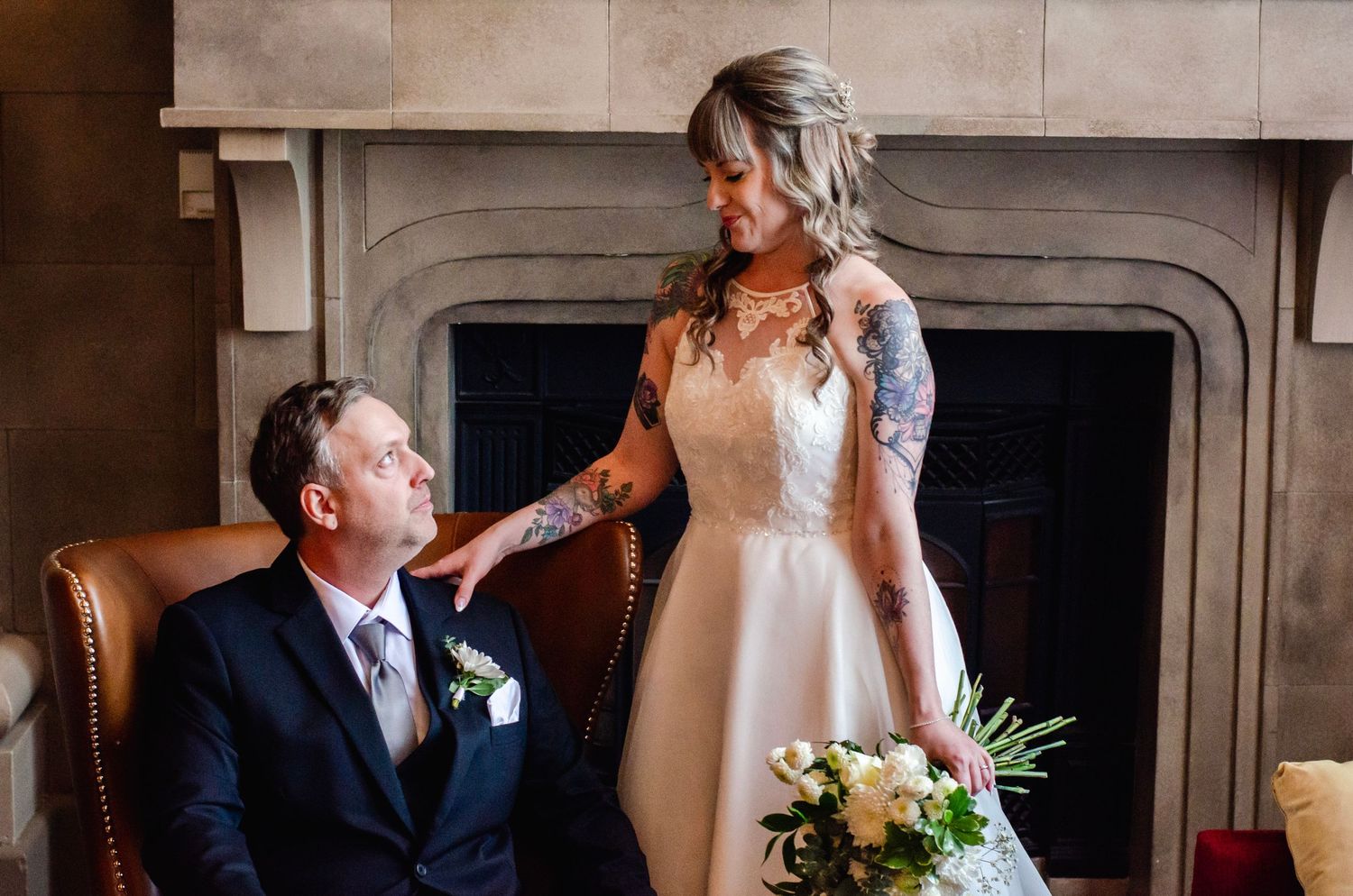 Bride in white gown with tattoos holds bouquet beside groom in navy suit seated by elegant fireplace.