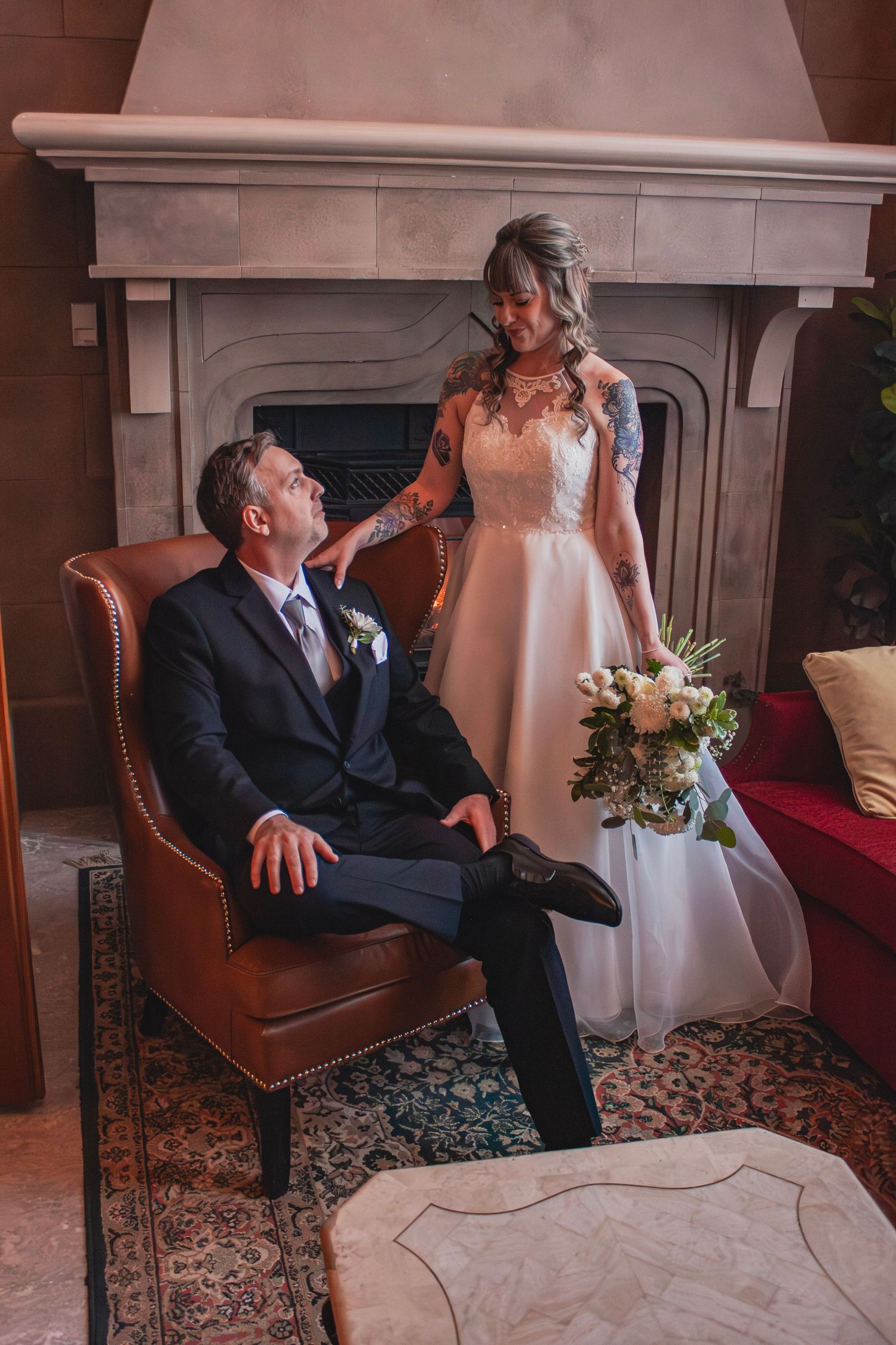 Bride with tattoos stands beside groom seated in leather chair near a fireplace, holding white bouquet.